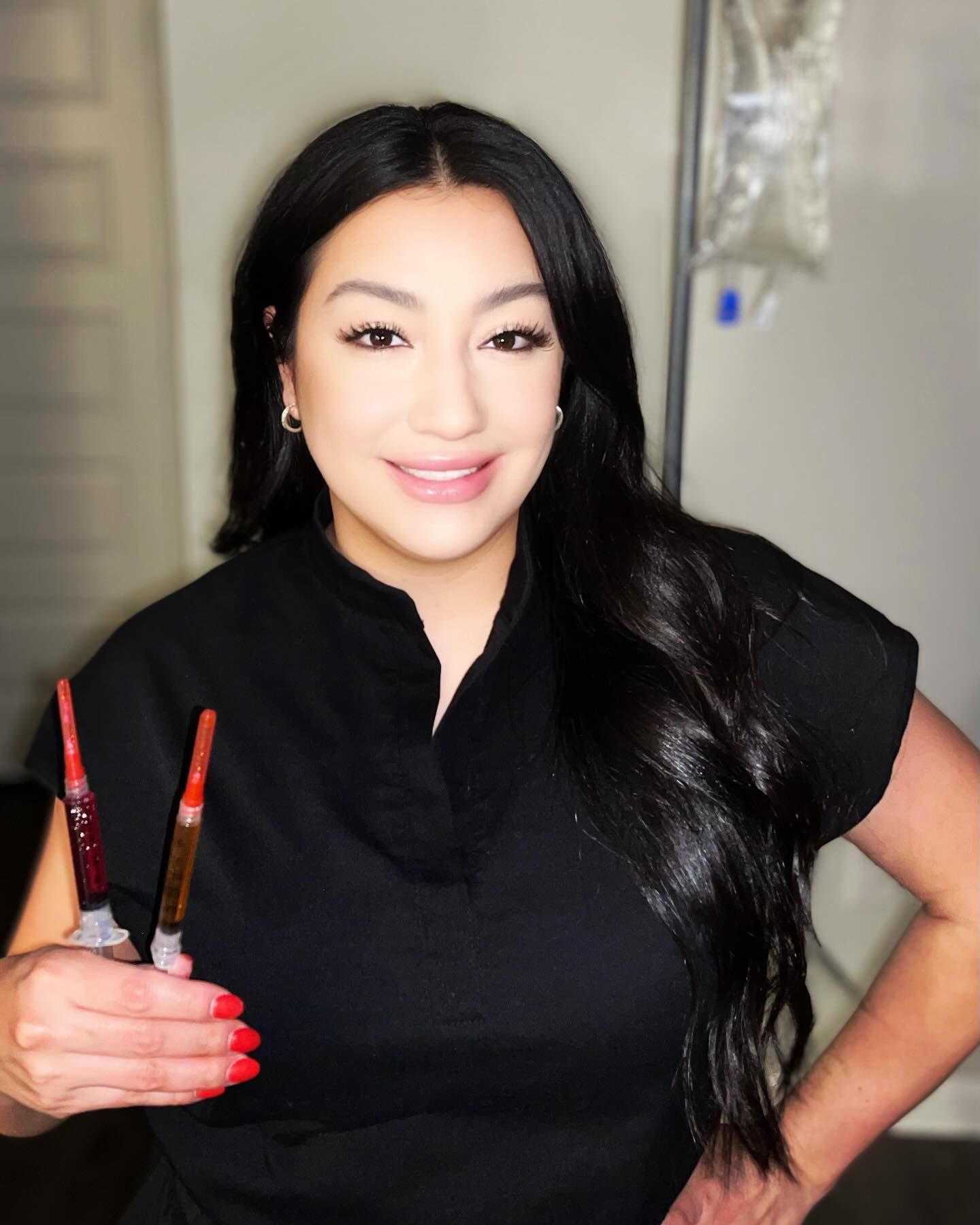 A smiling professional in black scrubs holds two syringes containing red and amber liquids in an indoor setting.