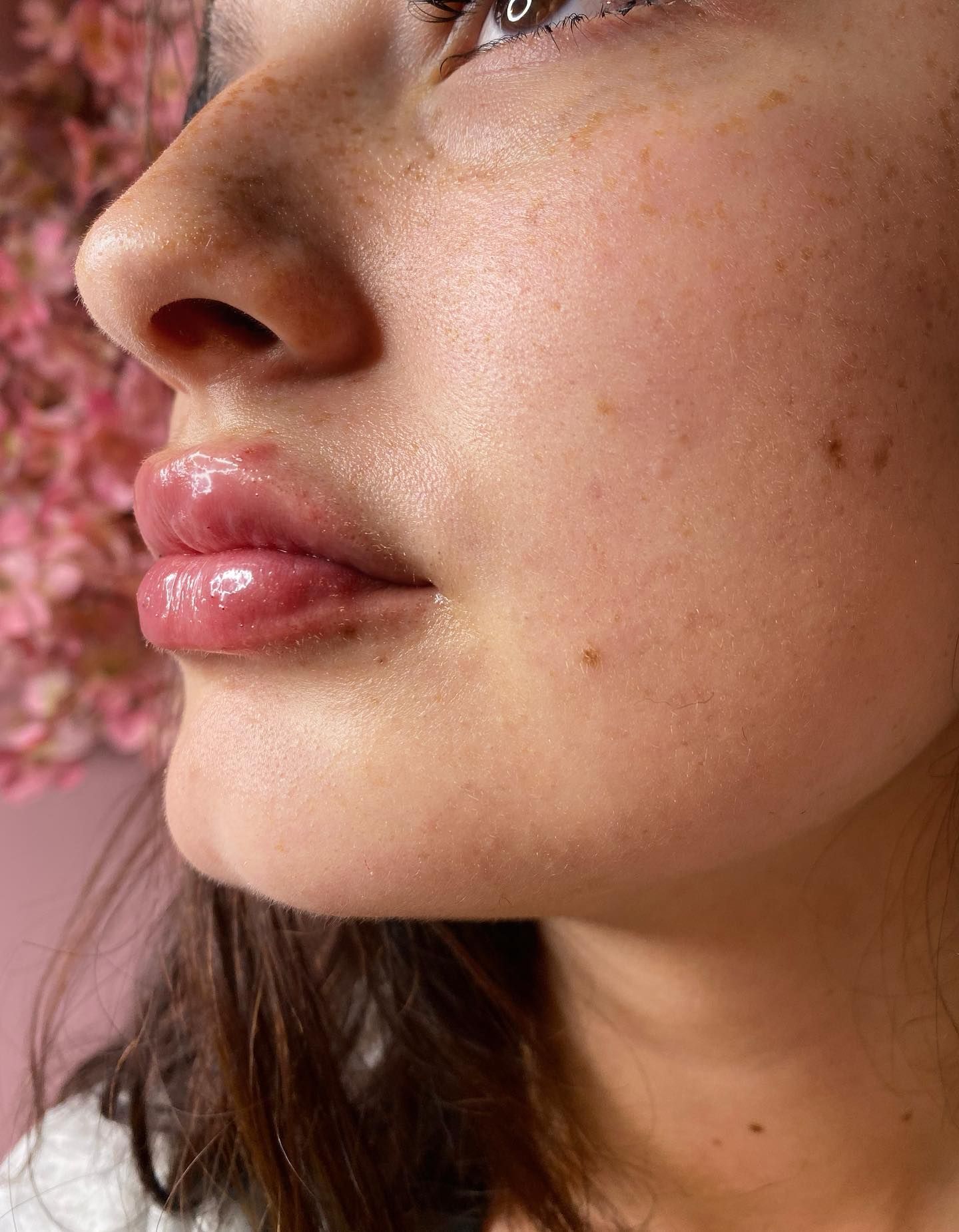 A close-up profile view of a face with glossy lips, light freckling, and natural skin texture against a pink background.