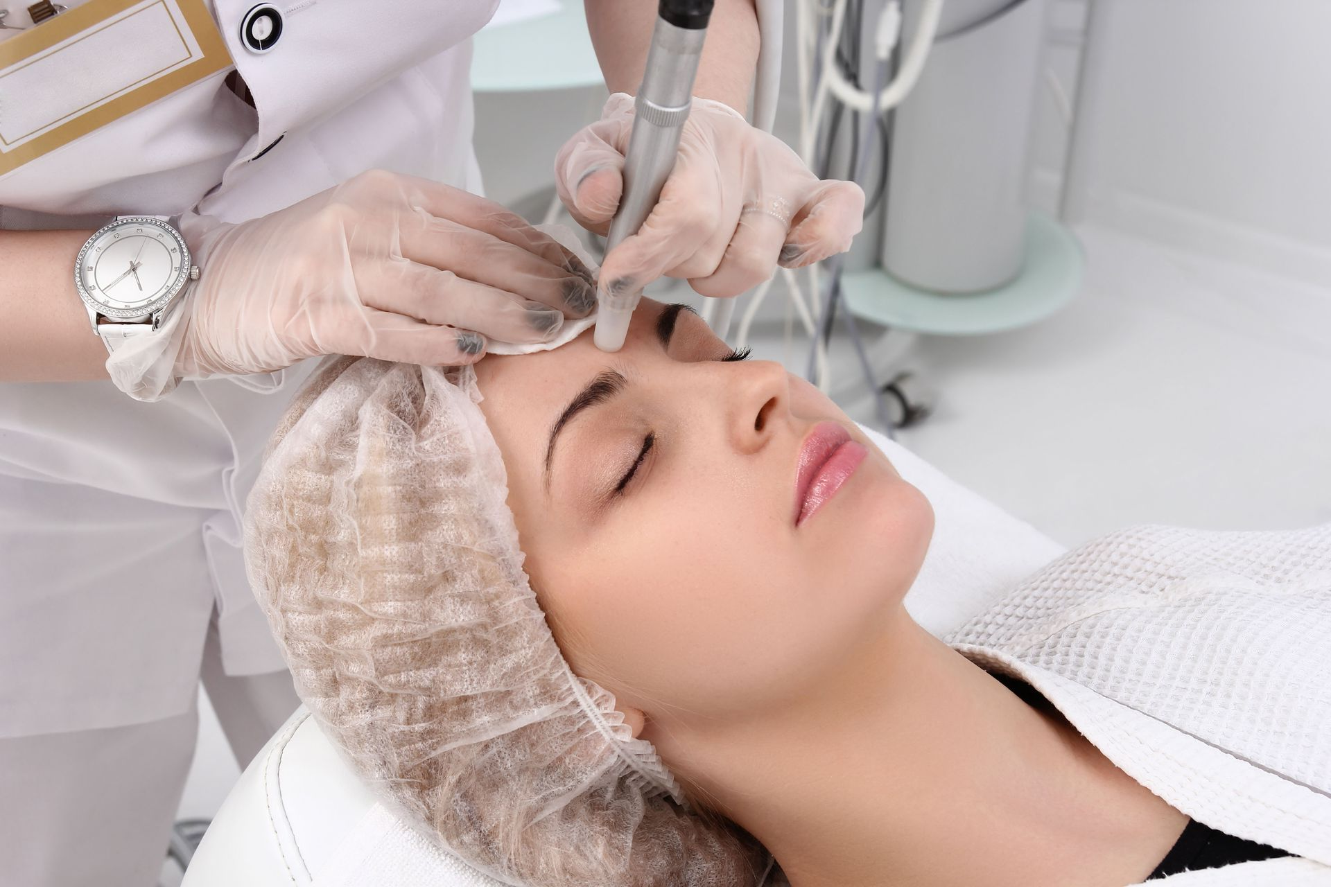 A professional performing a cosmetic skin treatment using a handheld device on a client&rsquo;s forehead in a clinic.