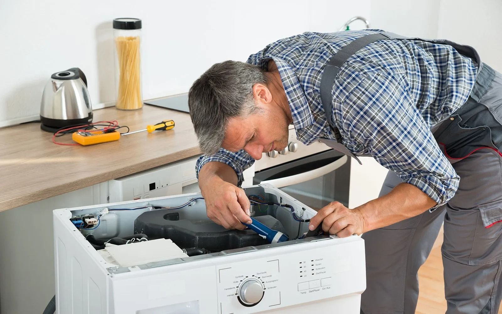 Man in overalls repairs a washing machine in a kitchen, using a screwdriver.
