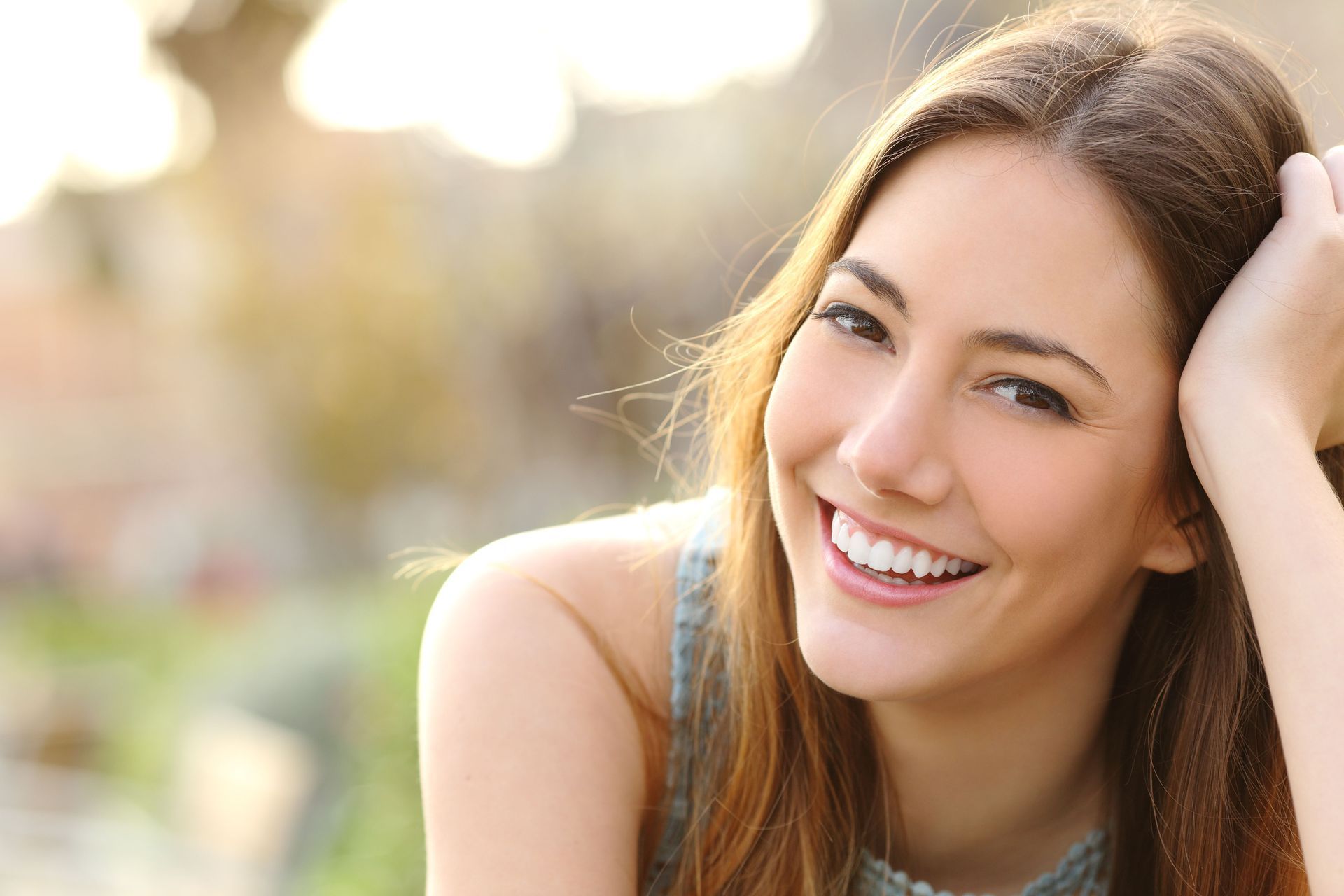 Woman with a wide smile, hand in hair, blurred outdoor background.