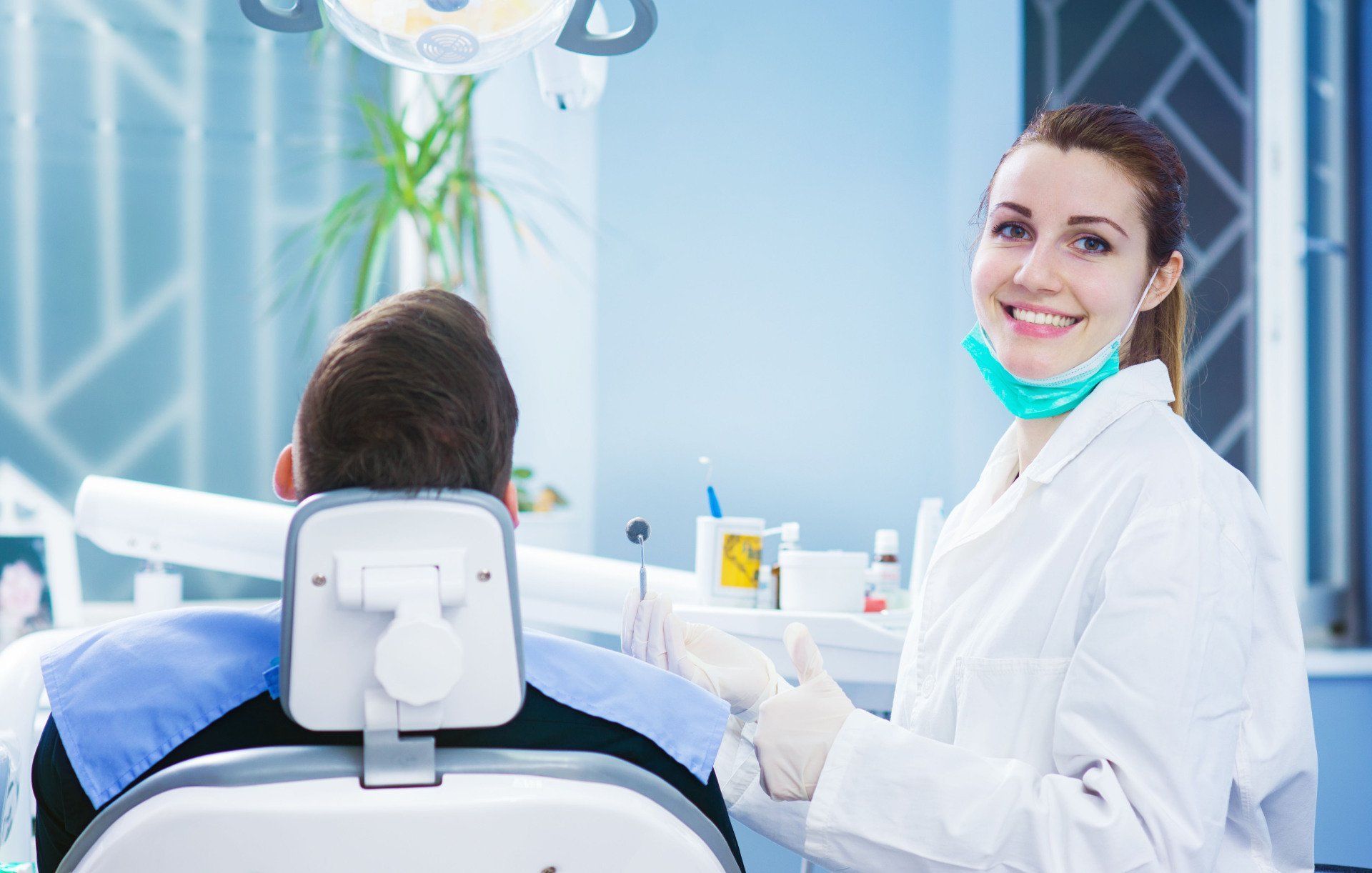 Dentist smiling at the camera, patient in chair. Clinic with dental equipment, white coat.