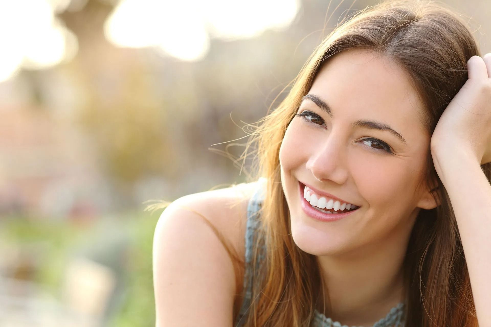 Woman smiling outdoors with a hand on her head, bright sunlight in background.