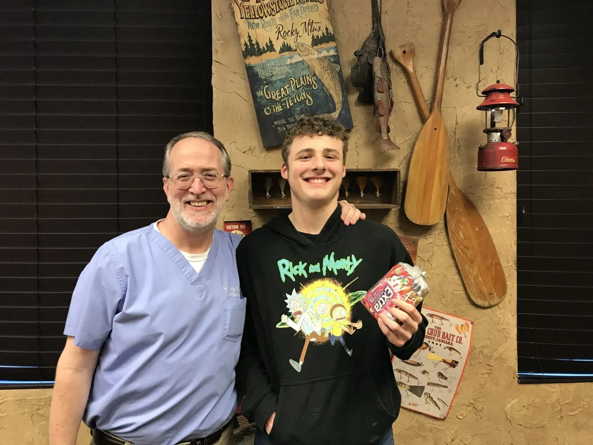 Man in scrubs stands with a teen holding a candy box, smiling. Rustic decor background.