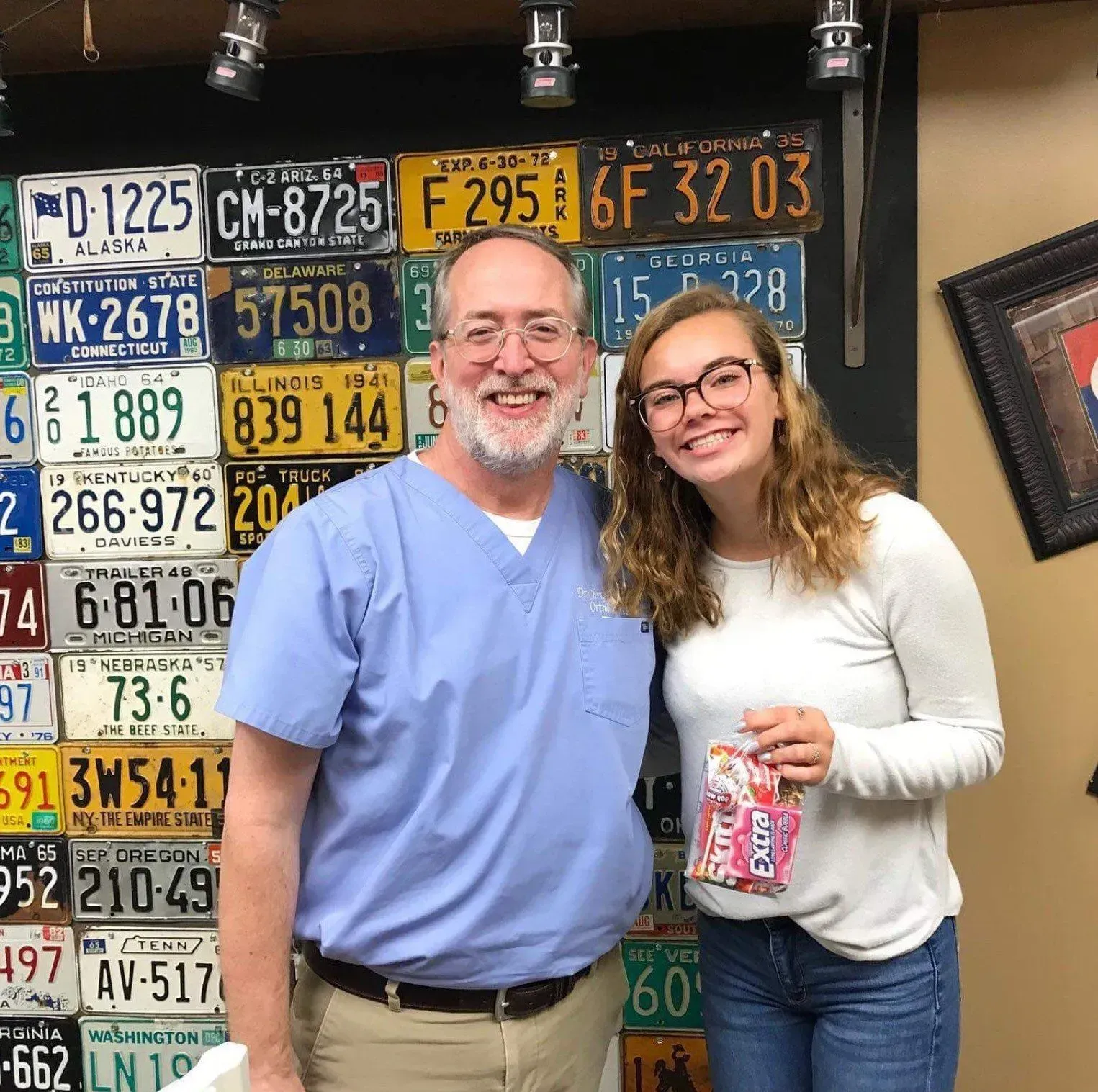 Man in scrubs and young woman smile, posing in front of license plates. The woman holds candy.