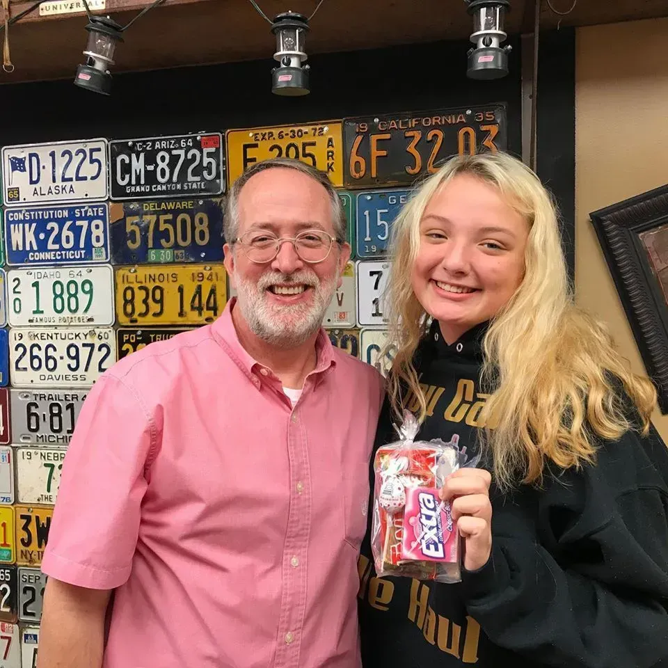 Man in pink shirt and young woman with blonde hair smiling, holding a bag of candy, in front of a license plate wall.