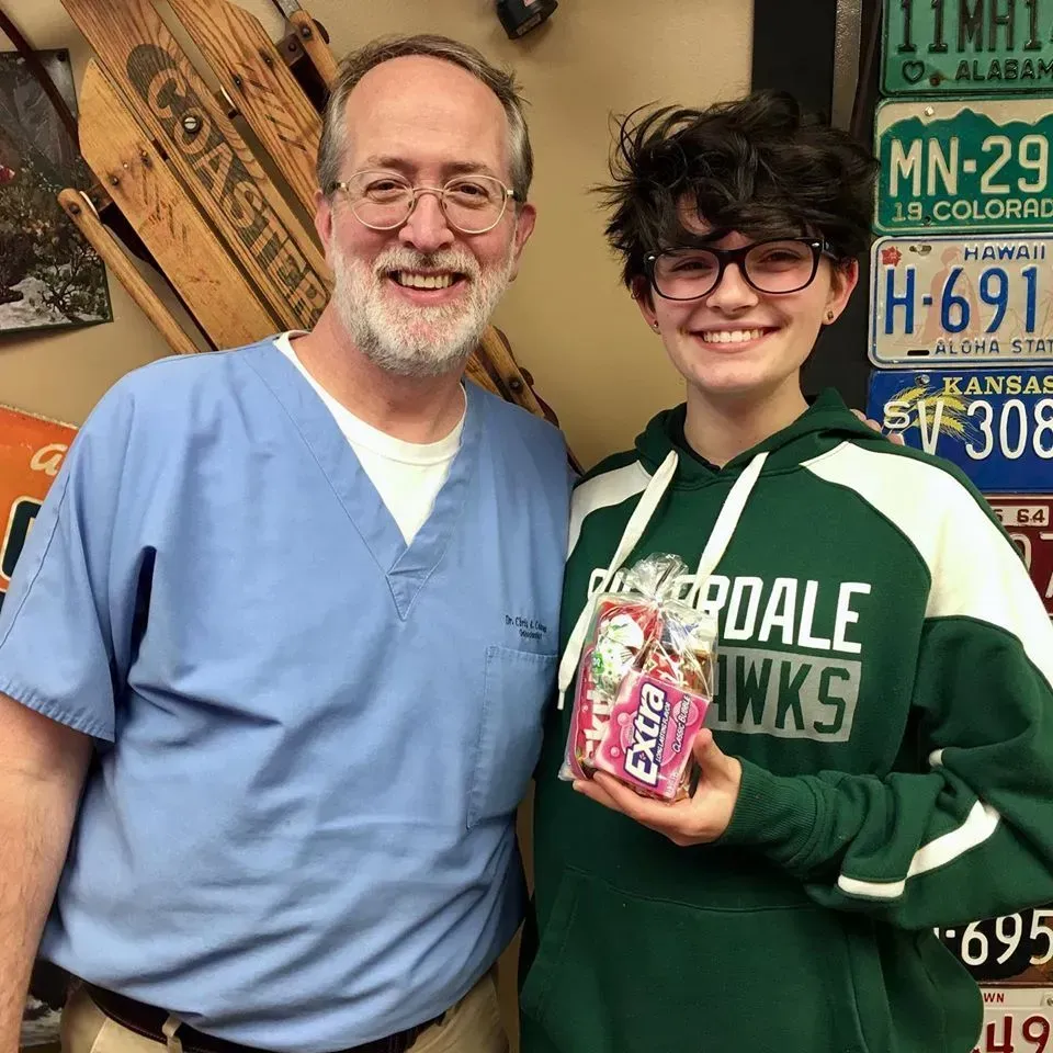 A smiling man in scrubs and a person in a green sweatshirt hold a pack of gum, license plates in background.