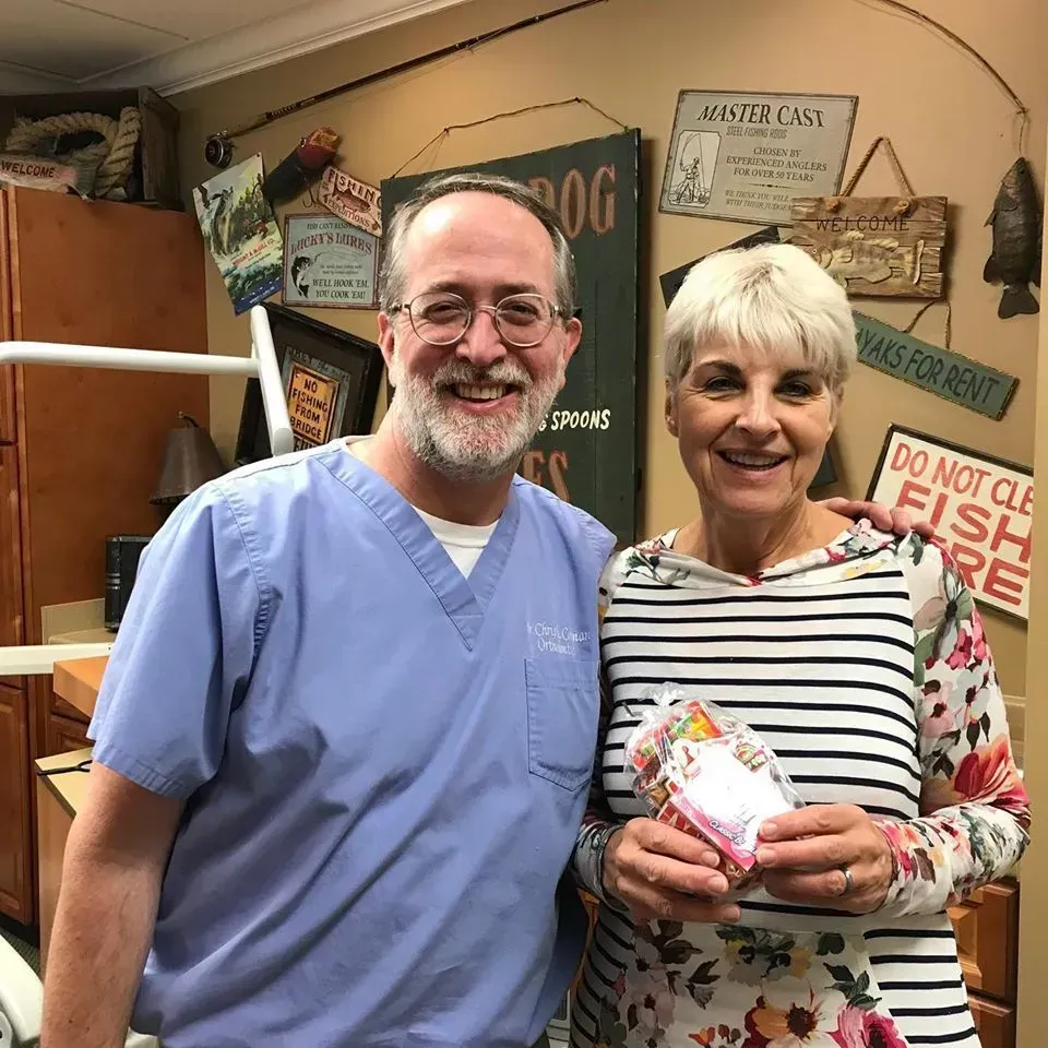 Man in scrubs and woman smiling, holding a gift. Beige walls, signs in background.