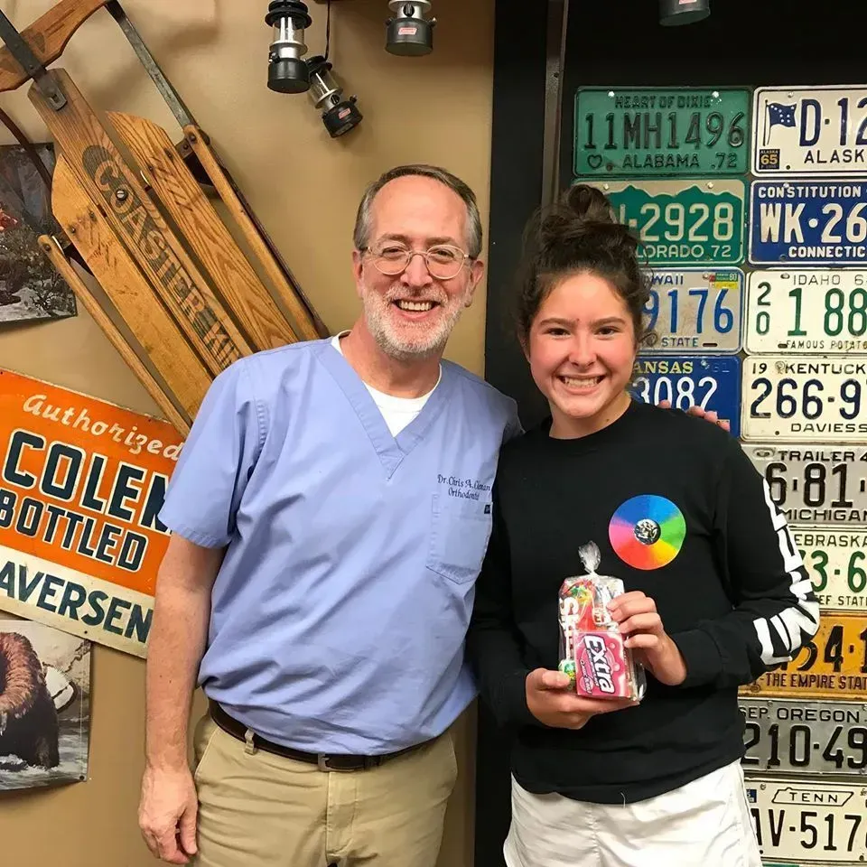A man in scrubs and a girl holding a treat pose in front of a wall with license plates and a vintage sign.