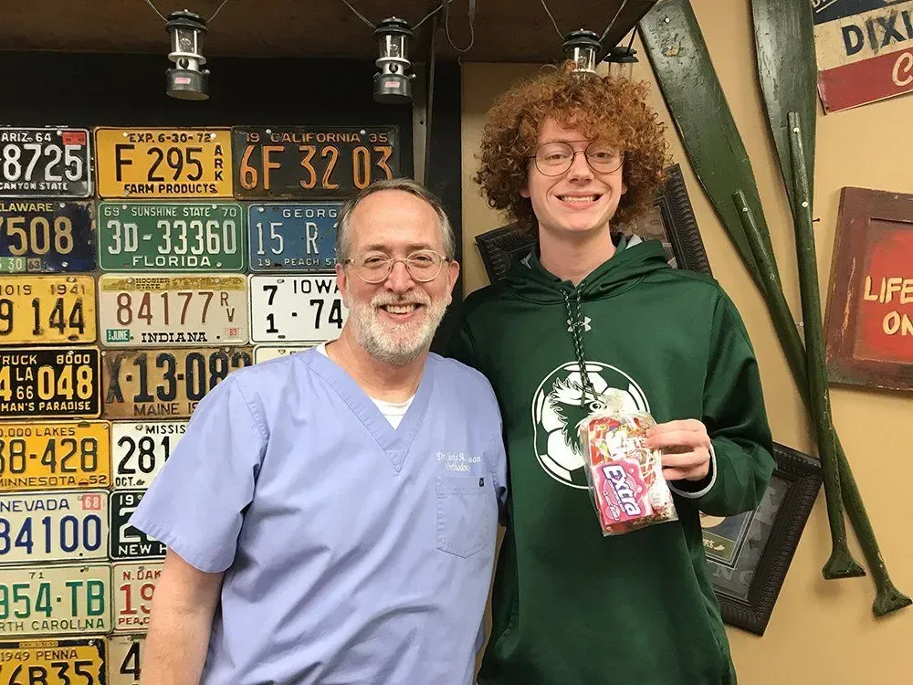 A man in scrubs and a young person pose smiling next to license plates and oars. The young person holds a snack.