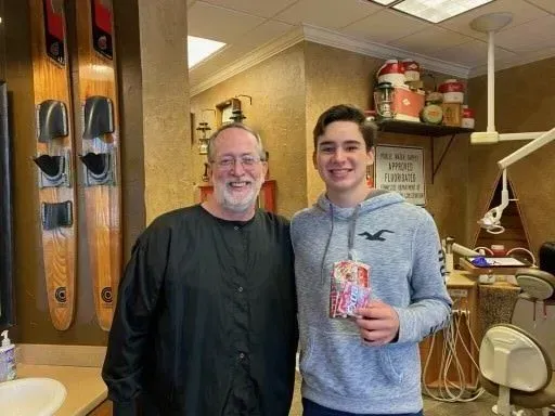 Man and teen in dentist's office, smiling. Teen holds small gift. Waterskis on wall.