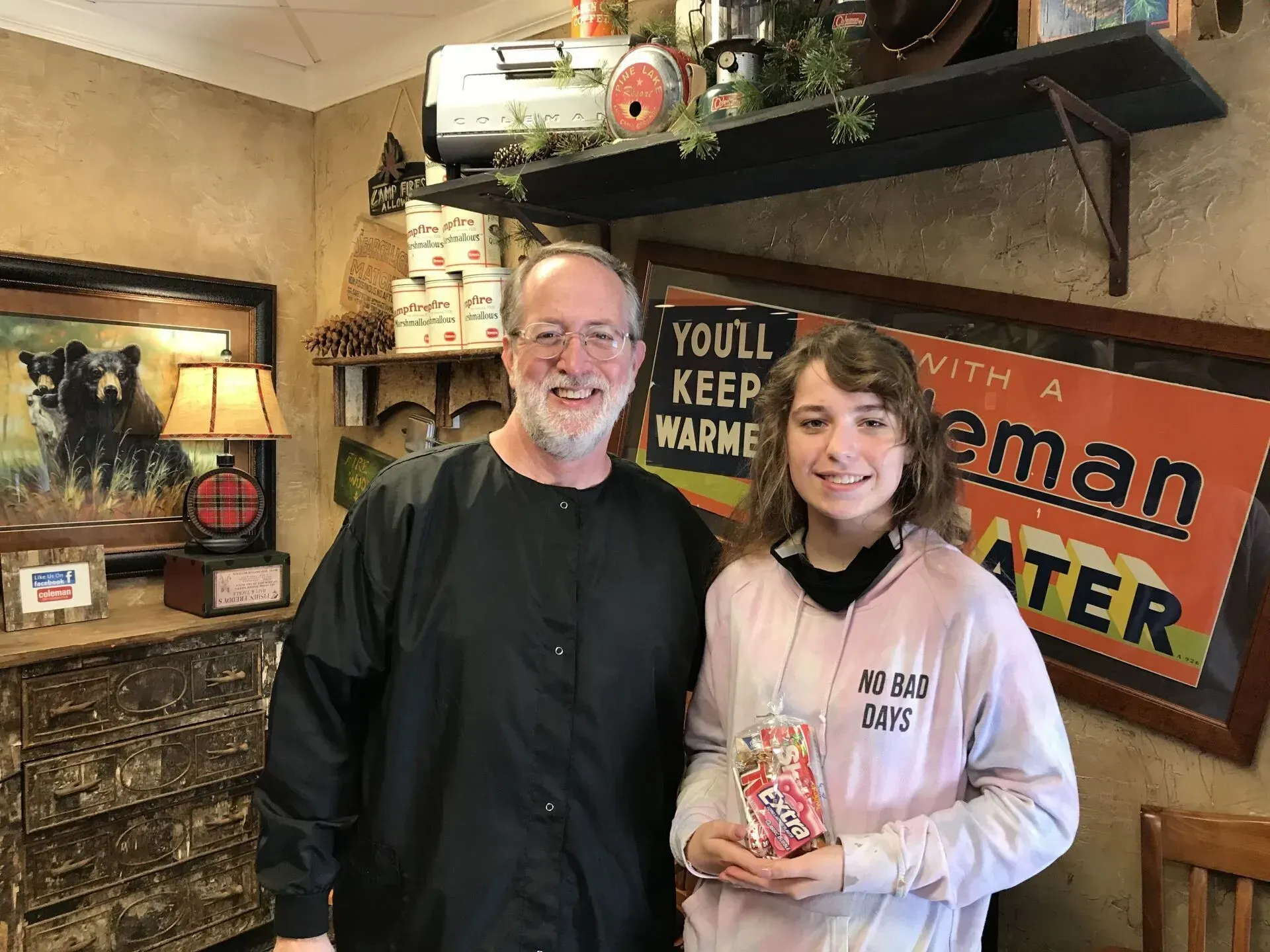 A man and a girl stand inside a shop. The girl holds a wrapped box, smiles. The man wears black. Rustic decor is visible.
