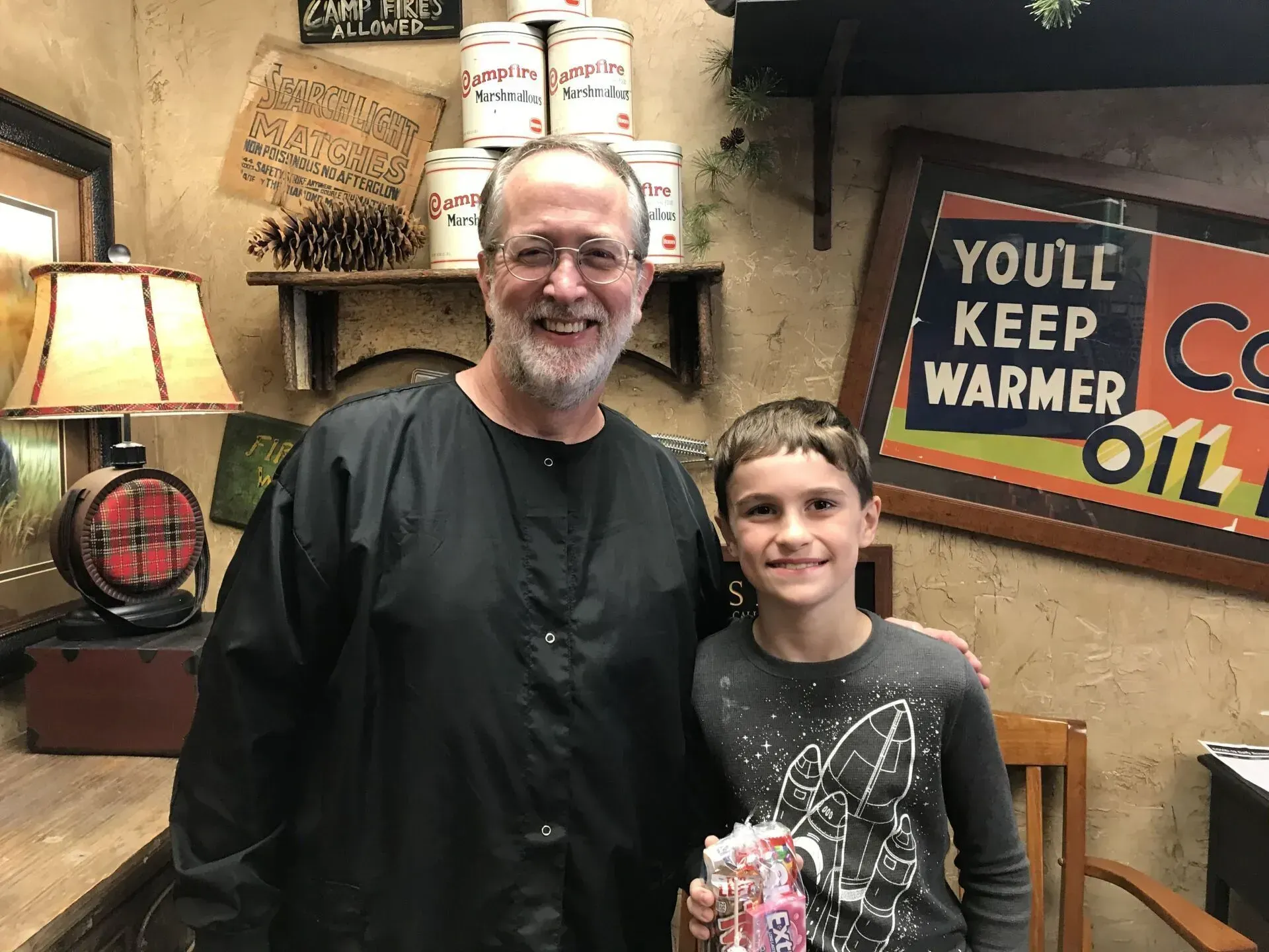 Man and boy smiling together in a barbershop. The man wears a barber's smock. Vintage decor is visible.
