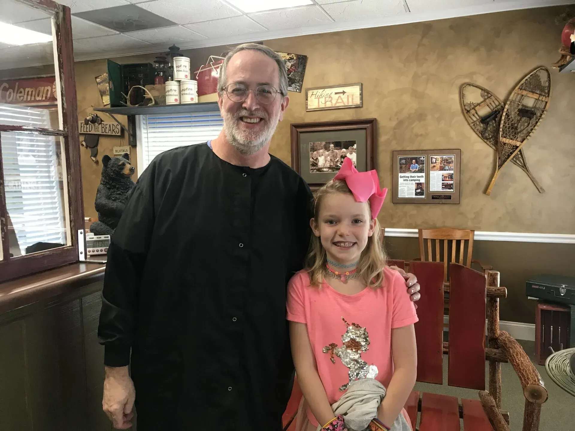 Man in black scrubs and young girl smiling in a dental office. Girl has a pink bow and shirt.