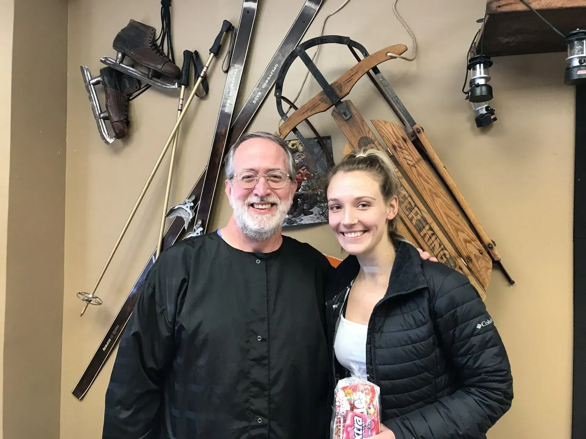 Man and young woman pose in front of vintage skis and sleds; woman holds a gift; beige wall.