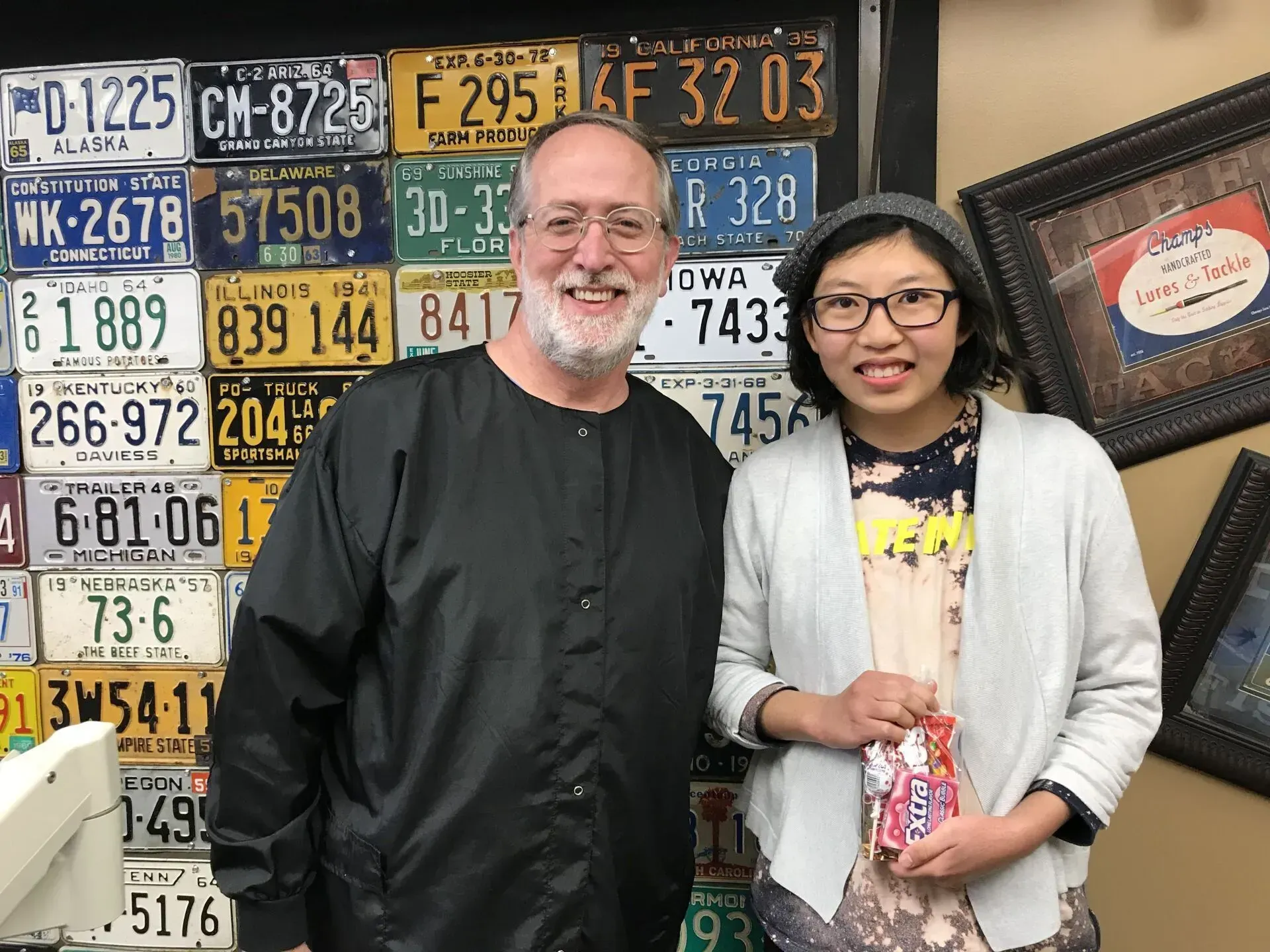 Two people smile in front of a wall of license plates. The person on the right holds candy.