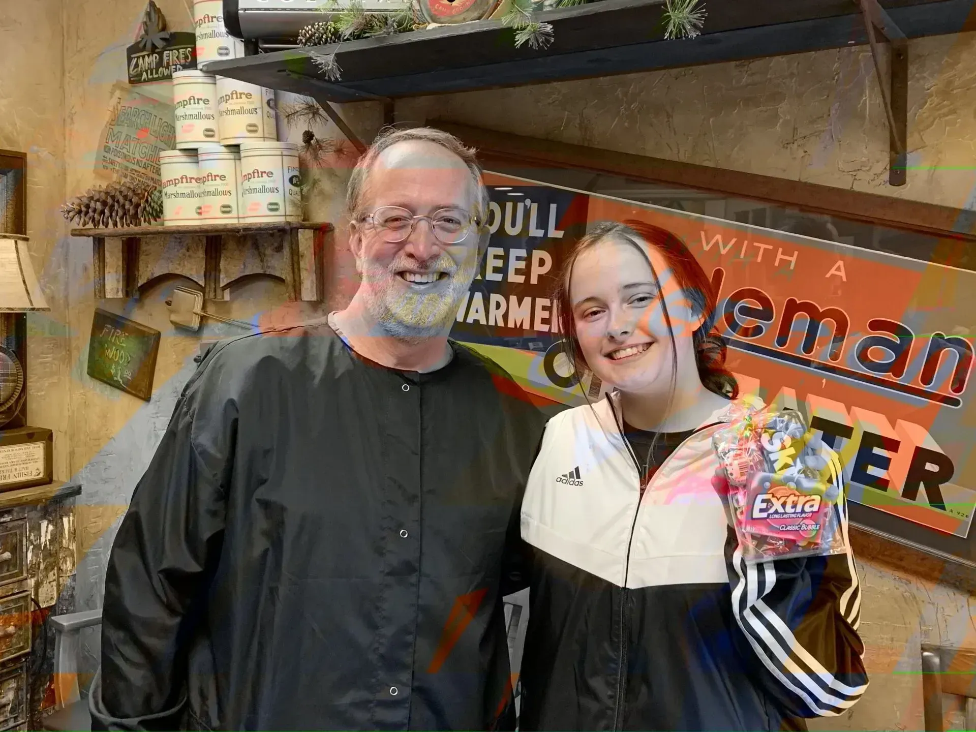 Man and girl smiling, holding gum, in front of a shop's colorful display, wooden sign, interior.