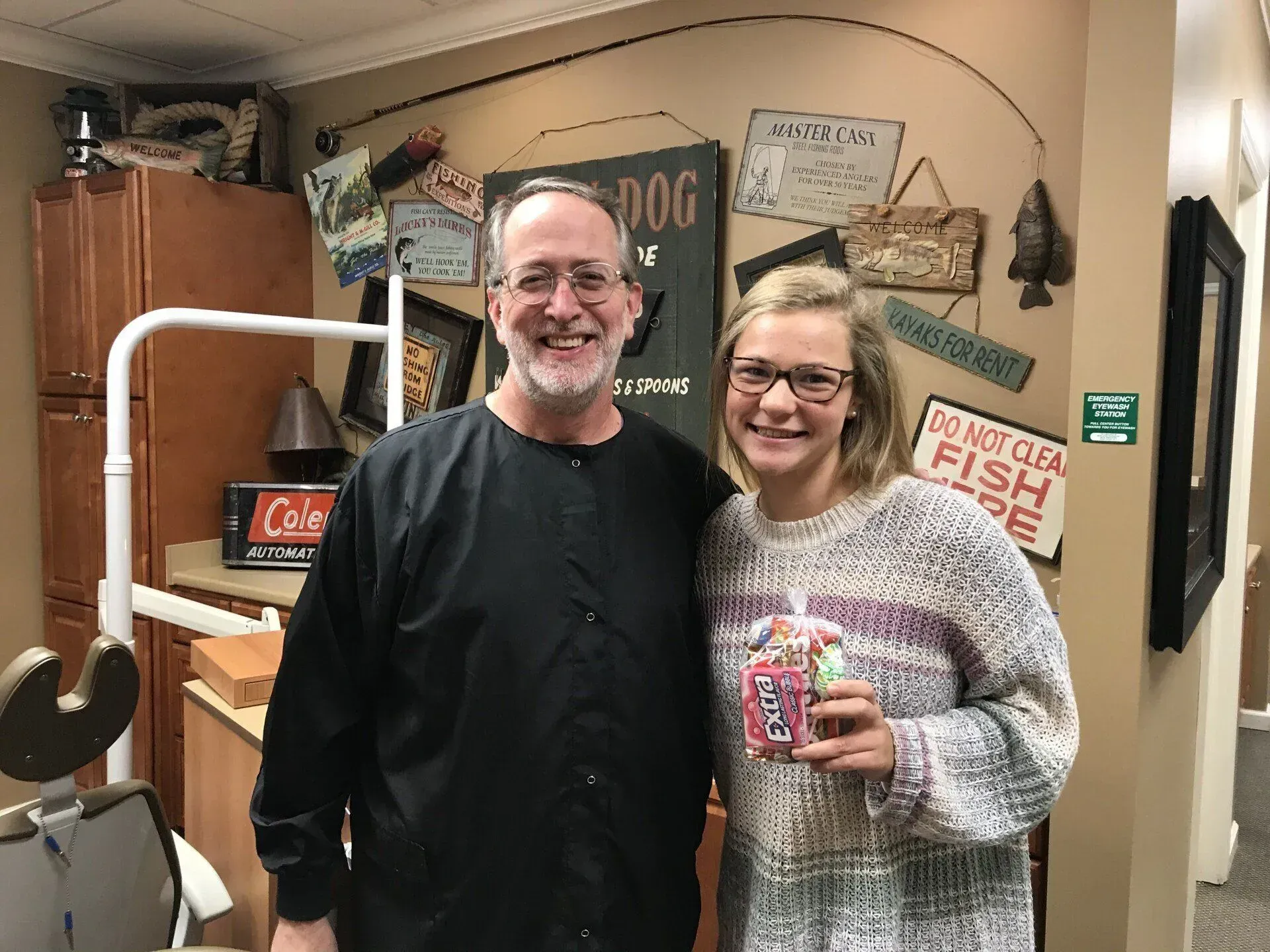 A smiling man and a young person pose together in a dental office. The young person holds a gift.