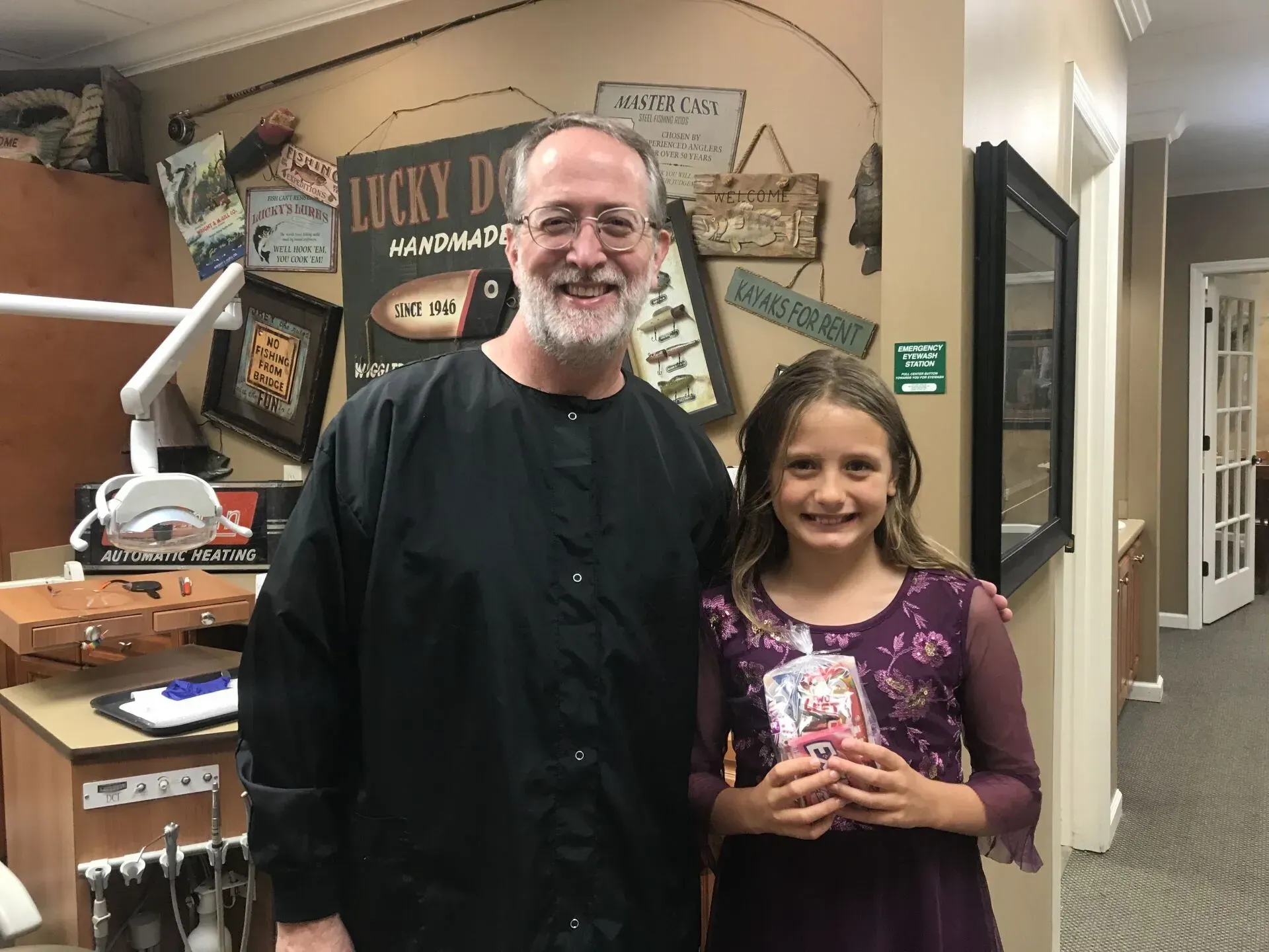 A dentist and a smiling girl posing in an office. Girl holds a treat. Background has wall decor.
