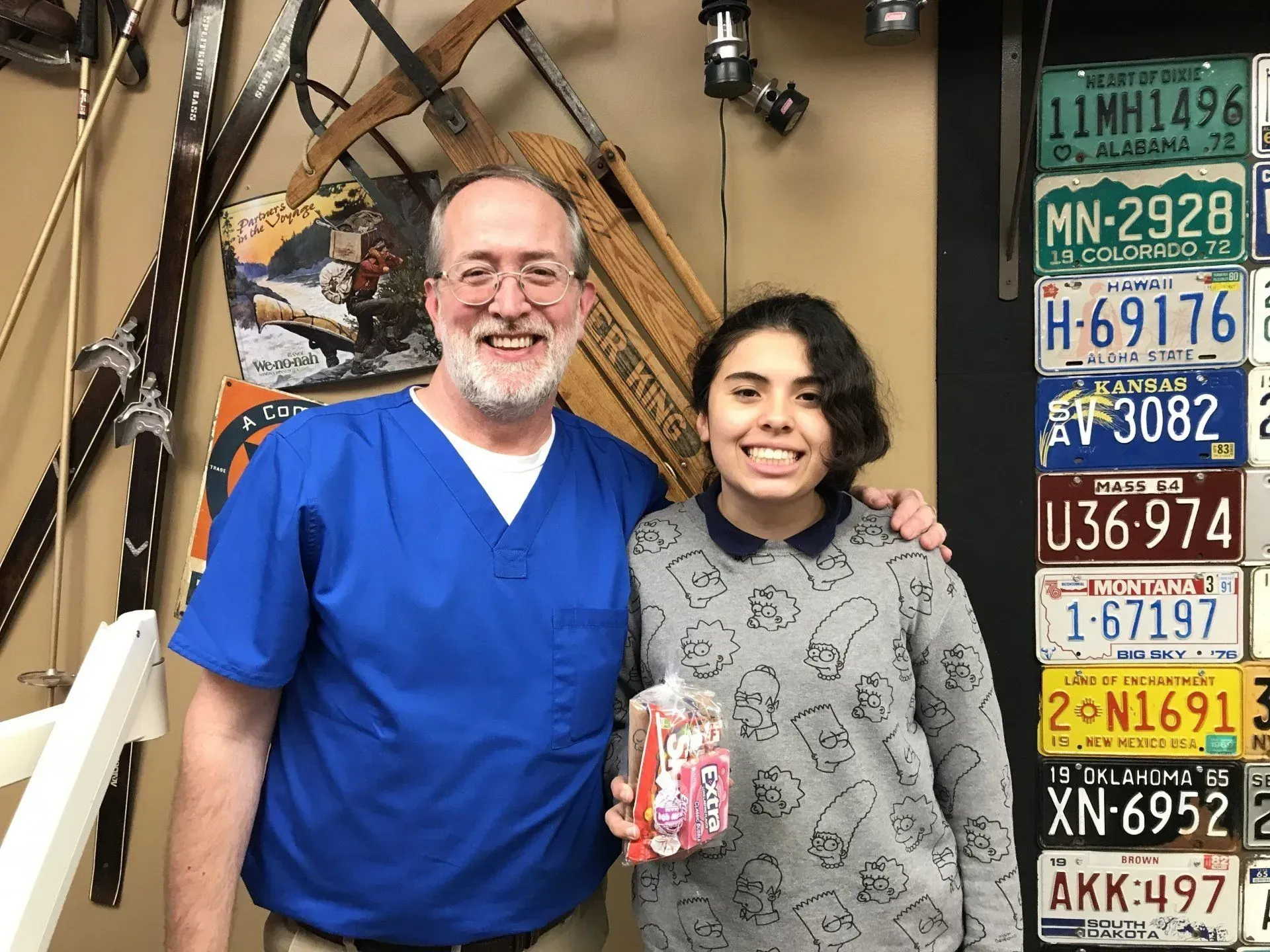 Man in blue scrubs and young person smiling, holding a gift, near license plates and swords.