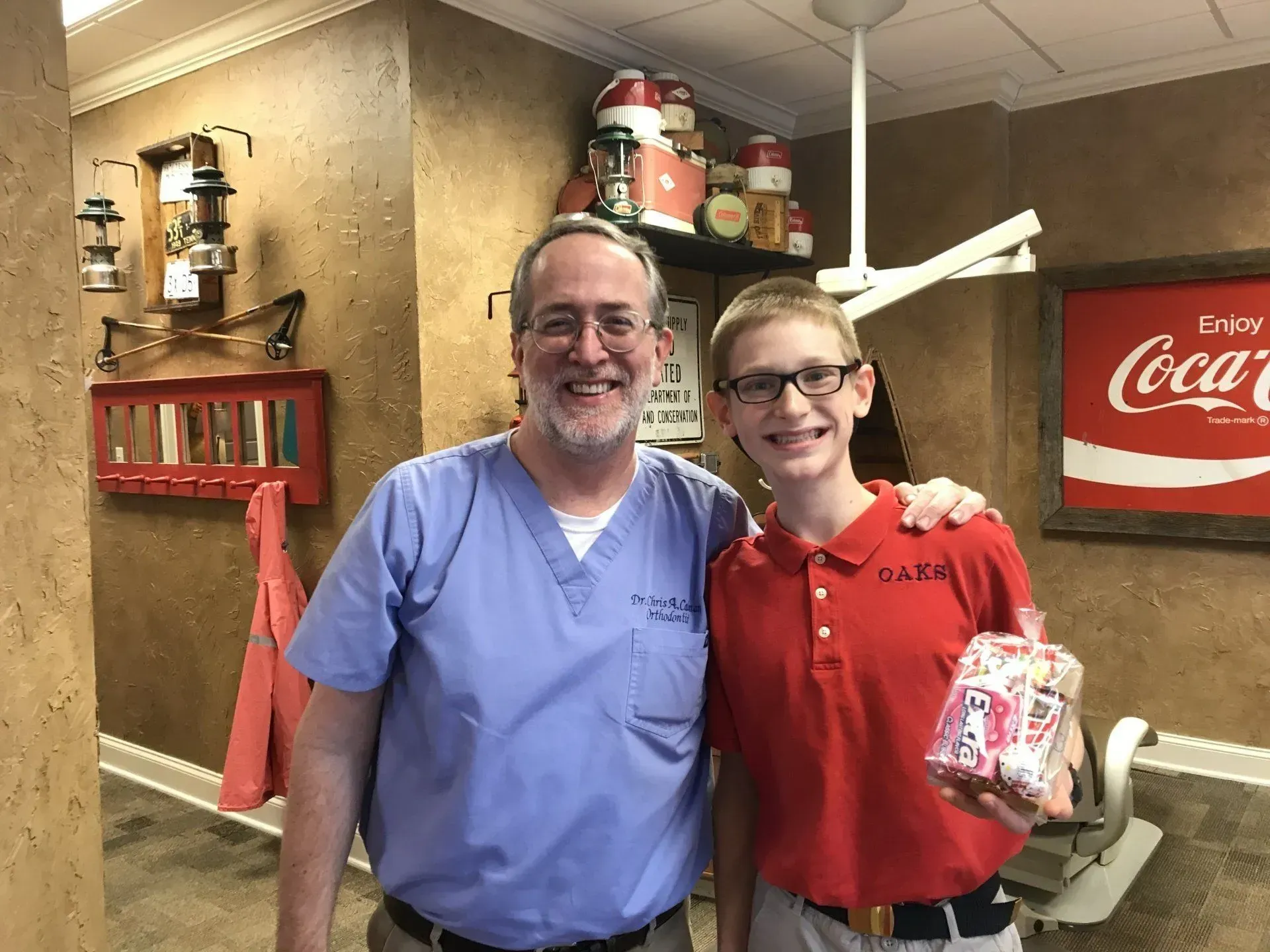 A dentist in blue scrubs smiles next to a boy in a red shirt holding a gift in a dentist's office.