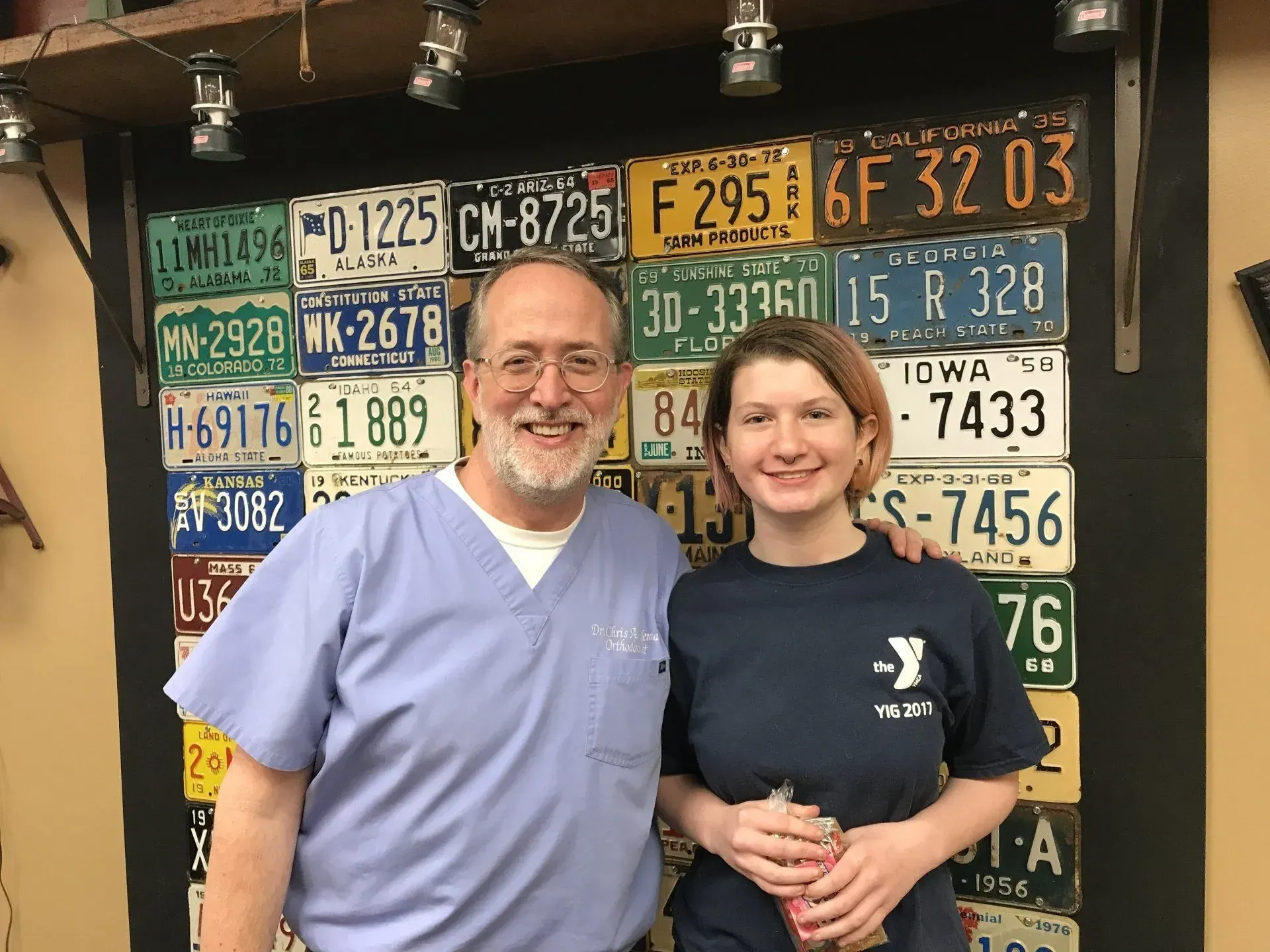 A man and woman smiling in front of a wall of license plates. The woman holds a cup.