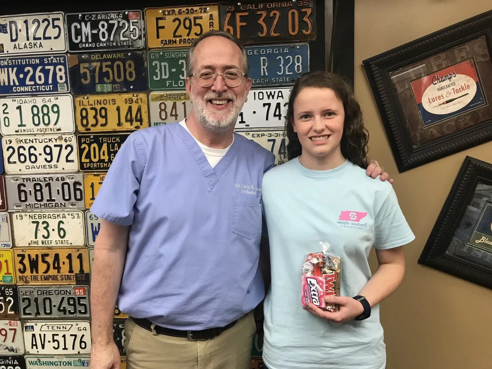 Man in scrubs and young woman smiling with gifts next to a wall of license plates.
