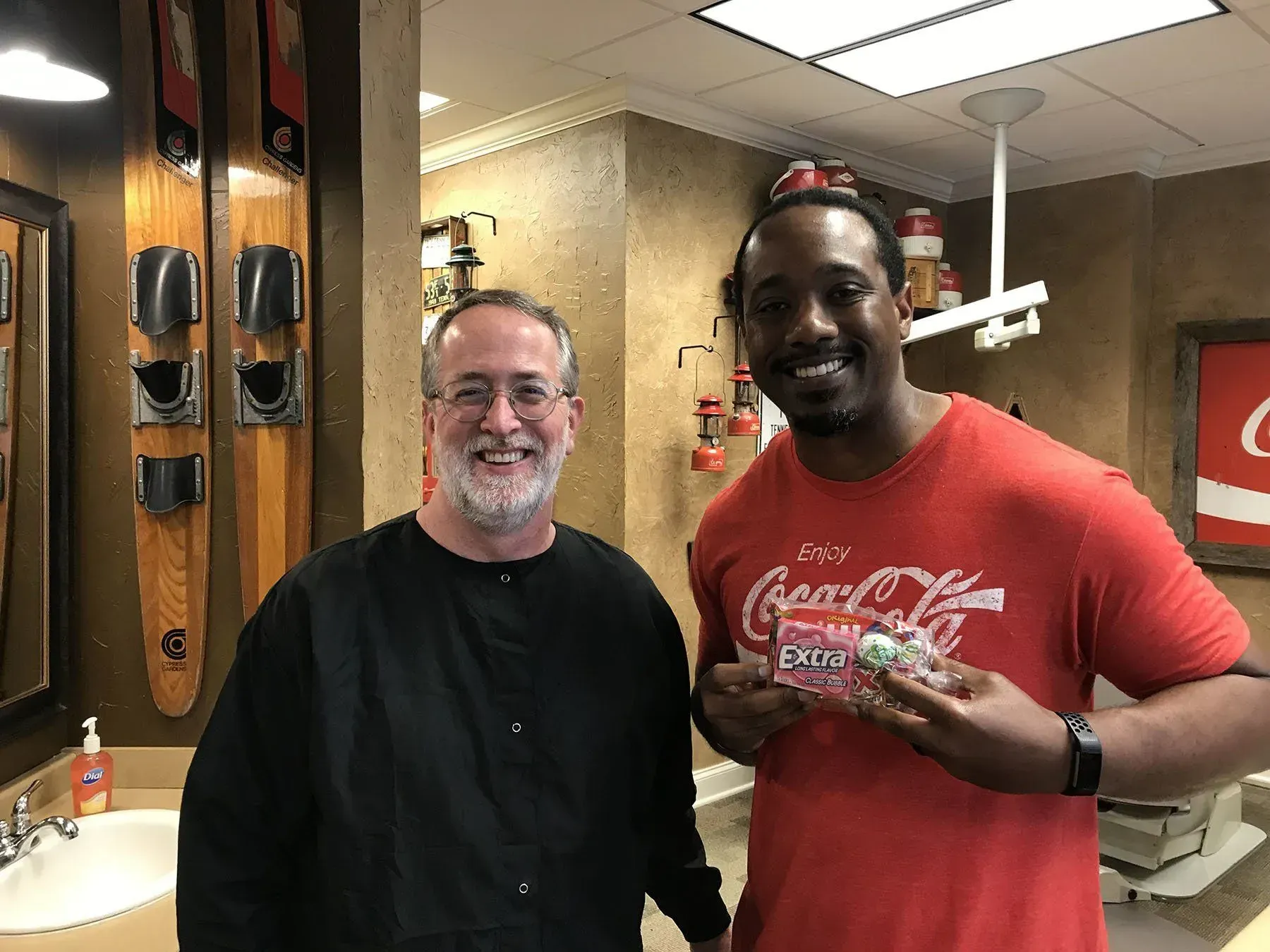 Two men smiling in a barbershop. One holds a pack of Excedrin. Wooden skis hang on the wall.