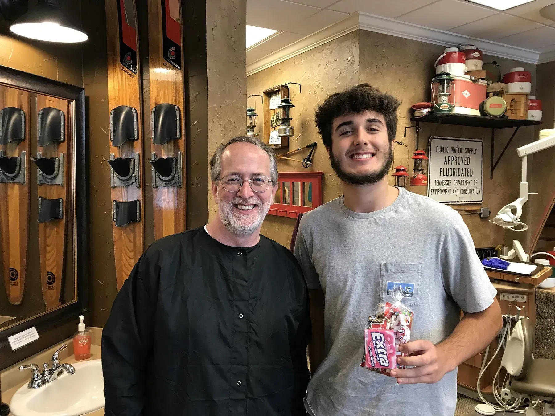 Man and young man smiling in a barbershop; the young man holds a gift bag. Water skis hang on the wall.