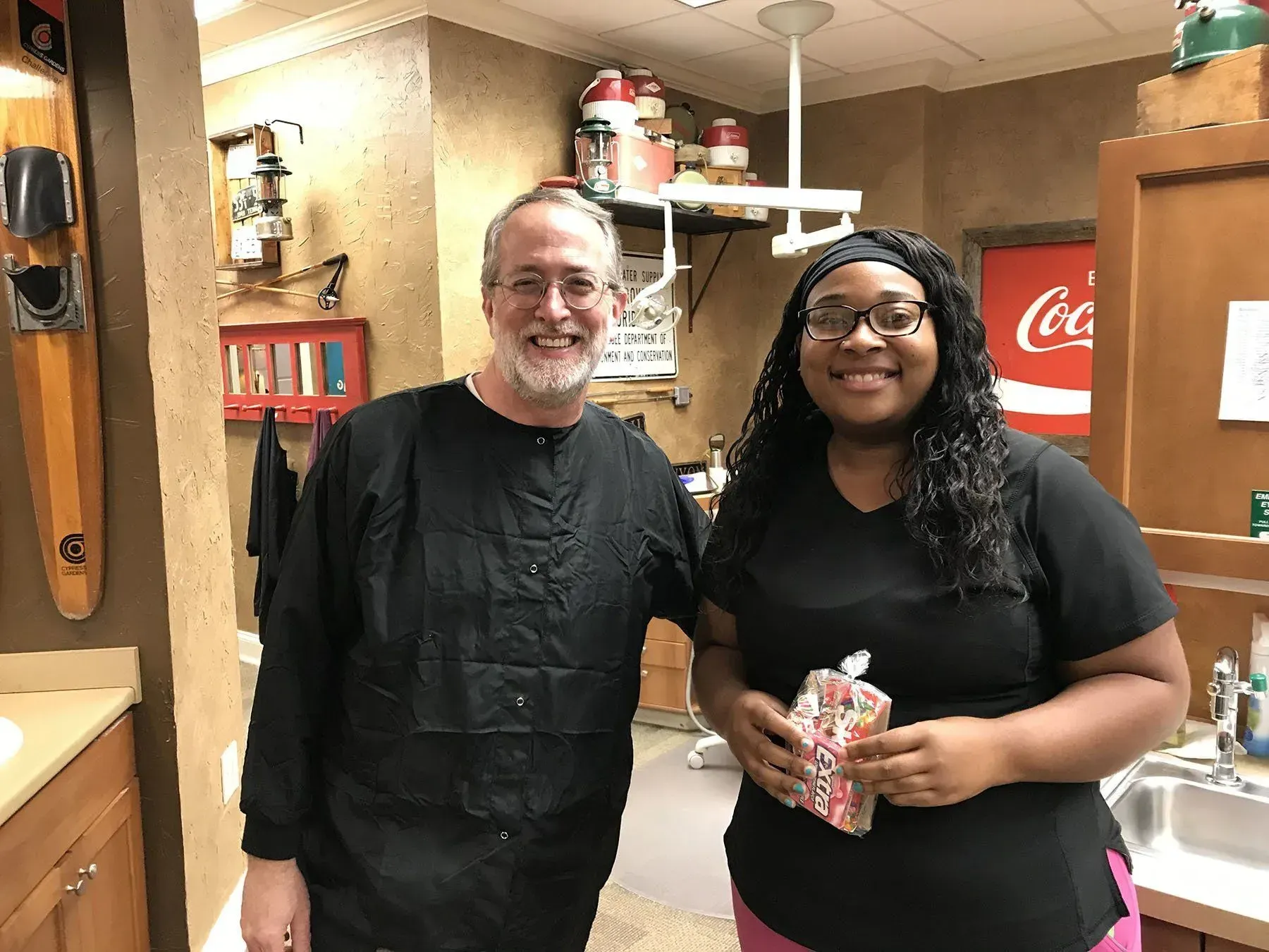 Two people in dental office, smiling. Man in black scrubs, woman in black top holding a treat. Red Coca-Cola sign.
