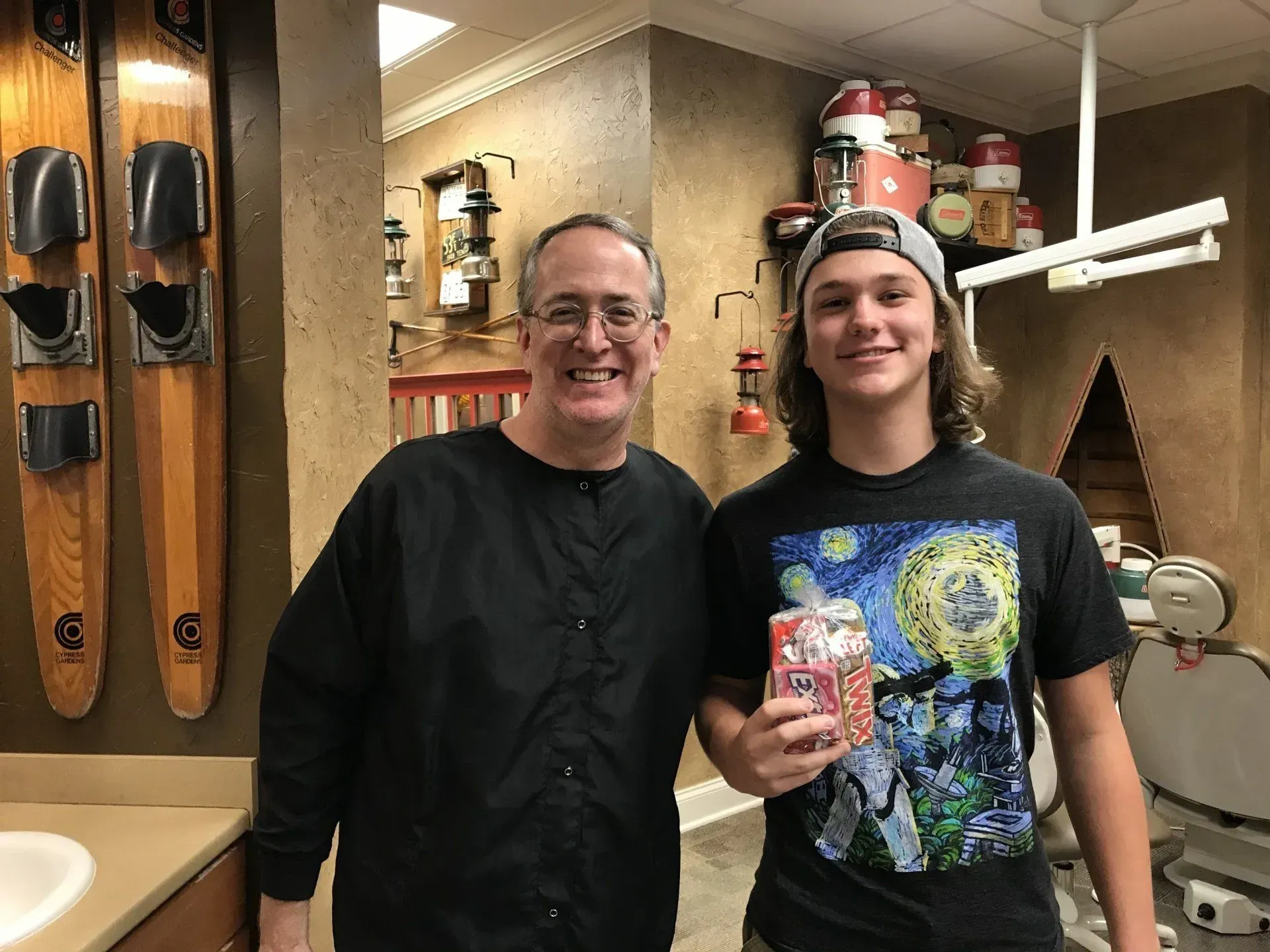 Man and teen in a dental office; both smiling. Teen holds small gift. Water skis and decor on walls.