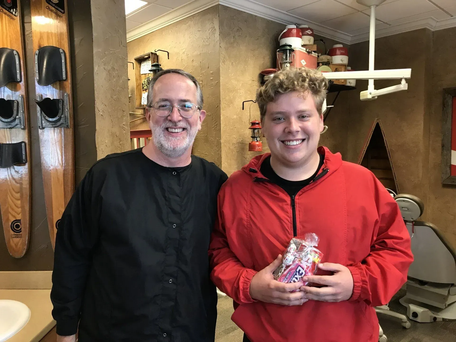 Man in black coat and young person in red jacket smiling, holding a gift, in a dental office.