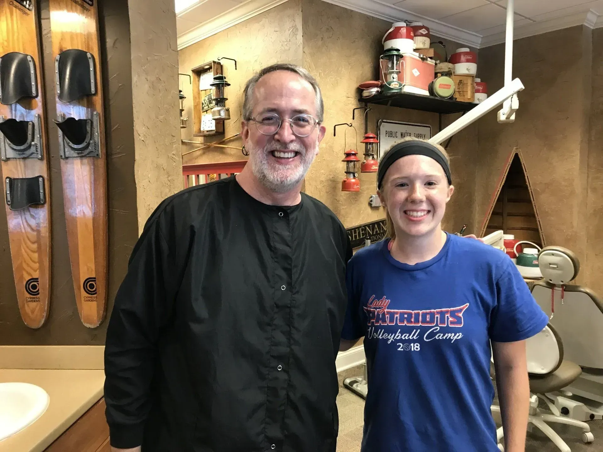 A smiling man and woman pose together in a dental office. The man is wearing glasses and a black jacket. 
