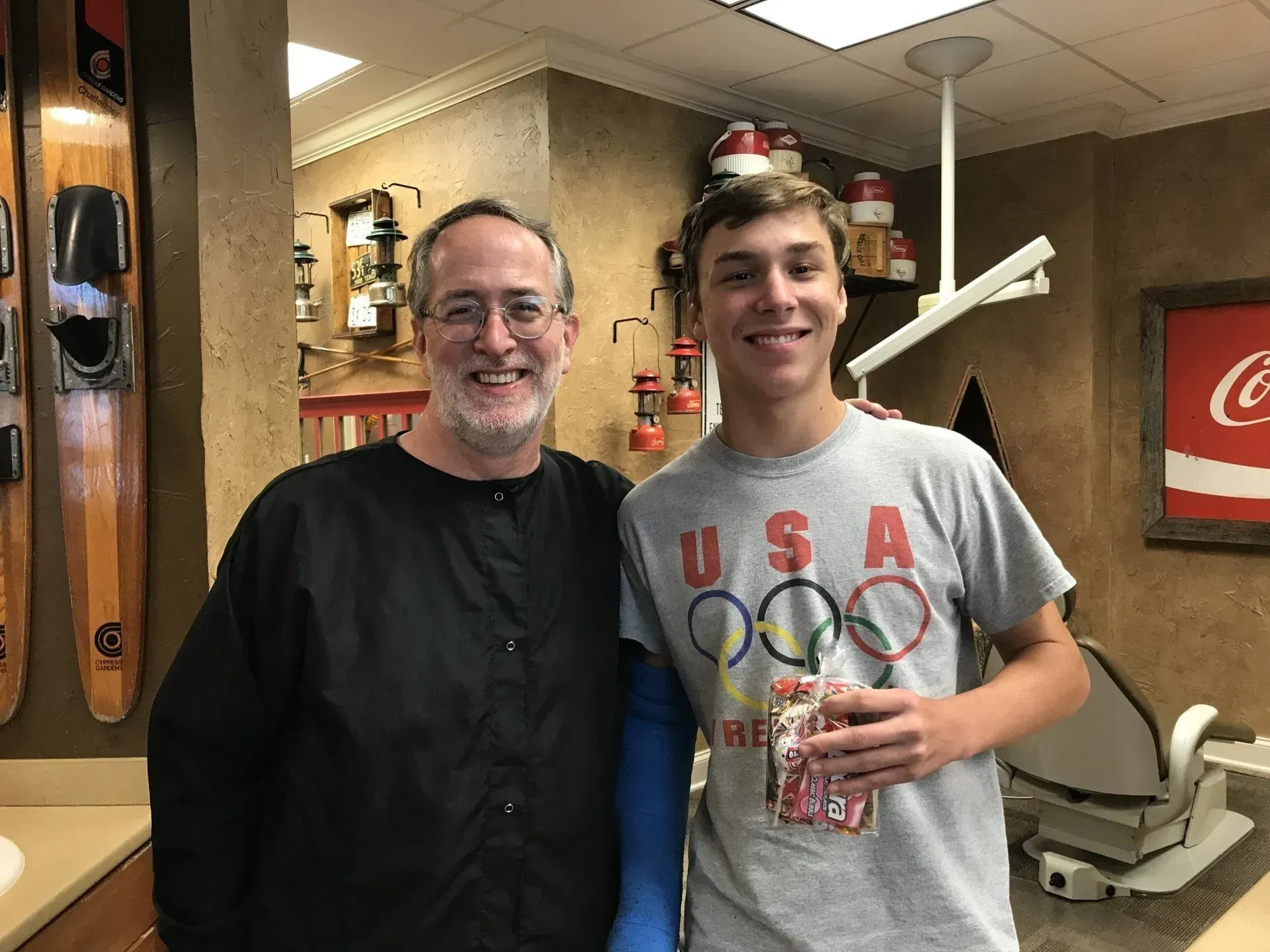 A man and a teen pose smiling. The teen holds a bag, the man wears a black coat, with ski decor on the wall behind them.