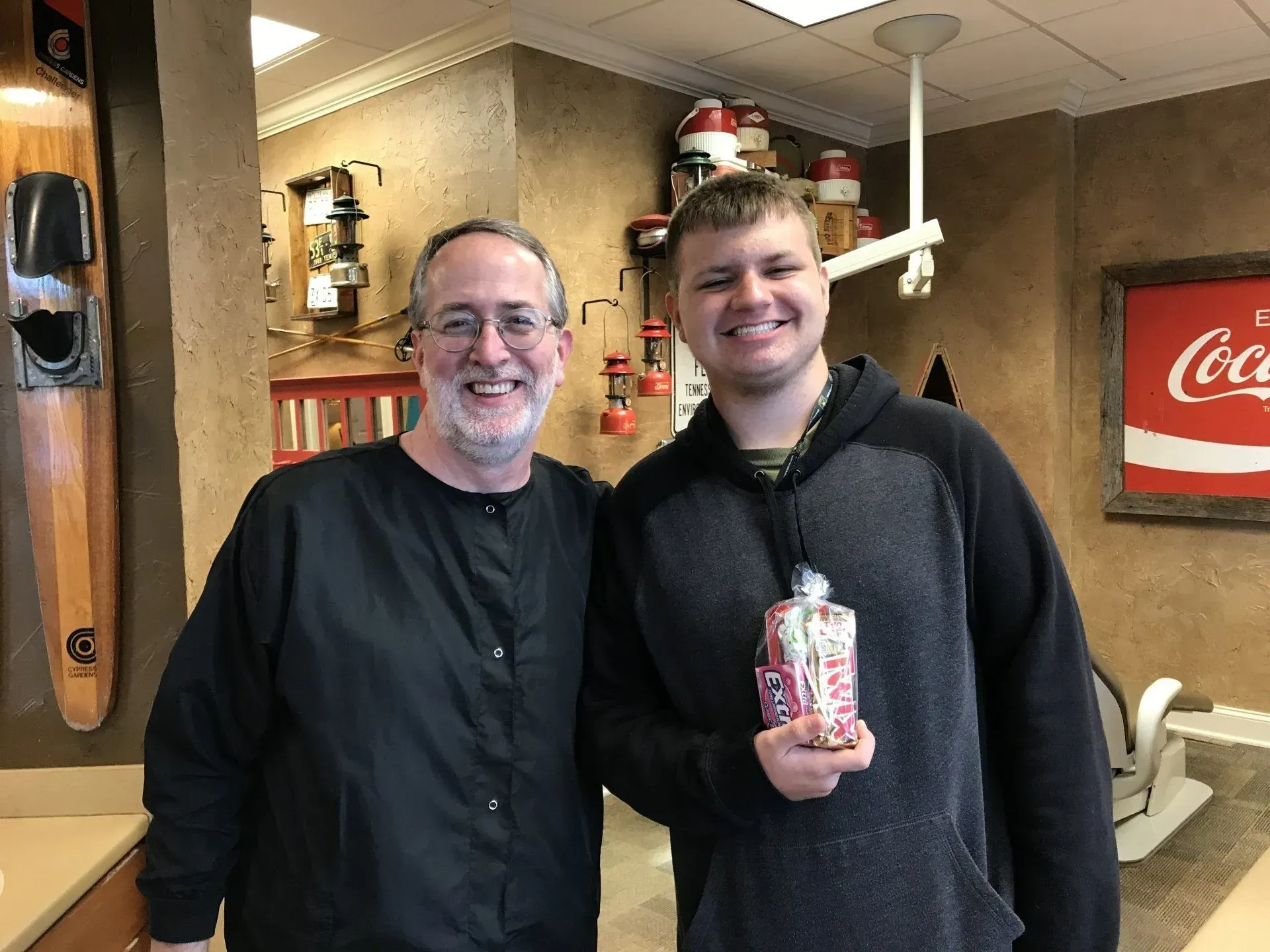 Two men smiling, one in black scrubs, the other holding a candy bag in a dental office. Coca-Cola sign in the background.