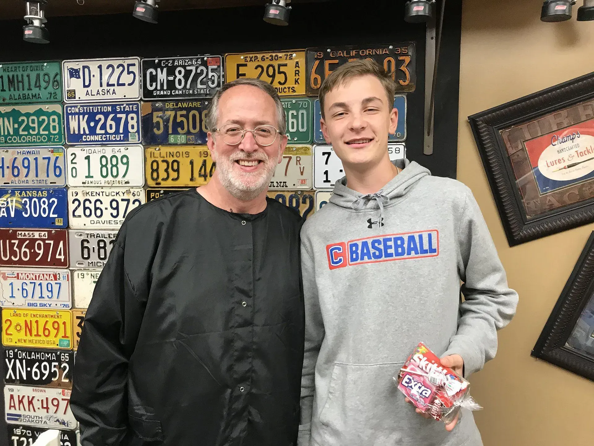 Two people pose smiling in front of a wall of license plates. The younger person holds candy.