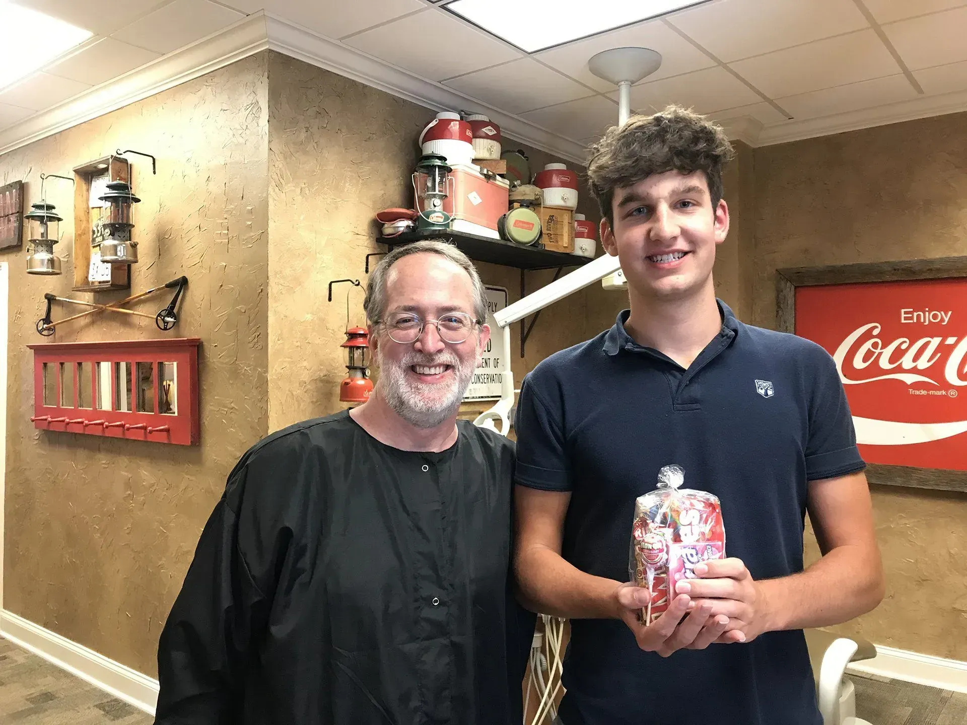 Man in black smock smiles with teen holding gift.  They stand in a room with vintage decor and Coca-Cola sign.