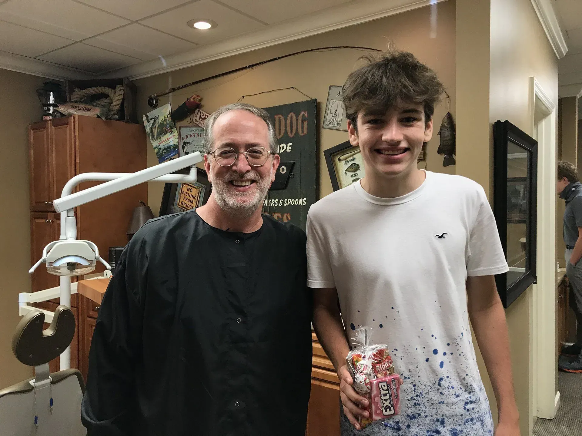 Orthodontist and patient smiling in office; dental equipment visible. Patient holds a drink.