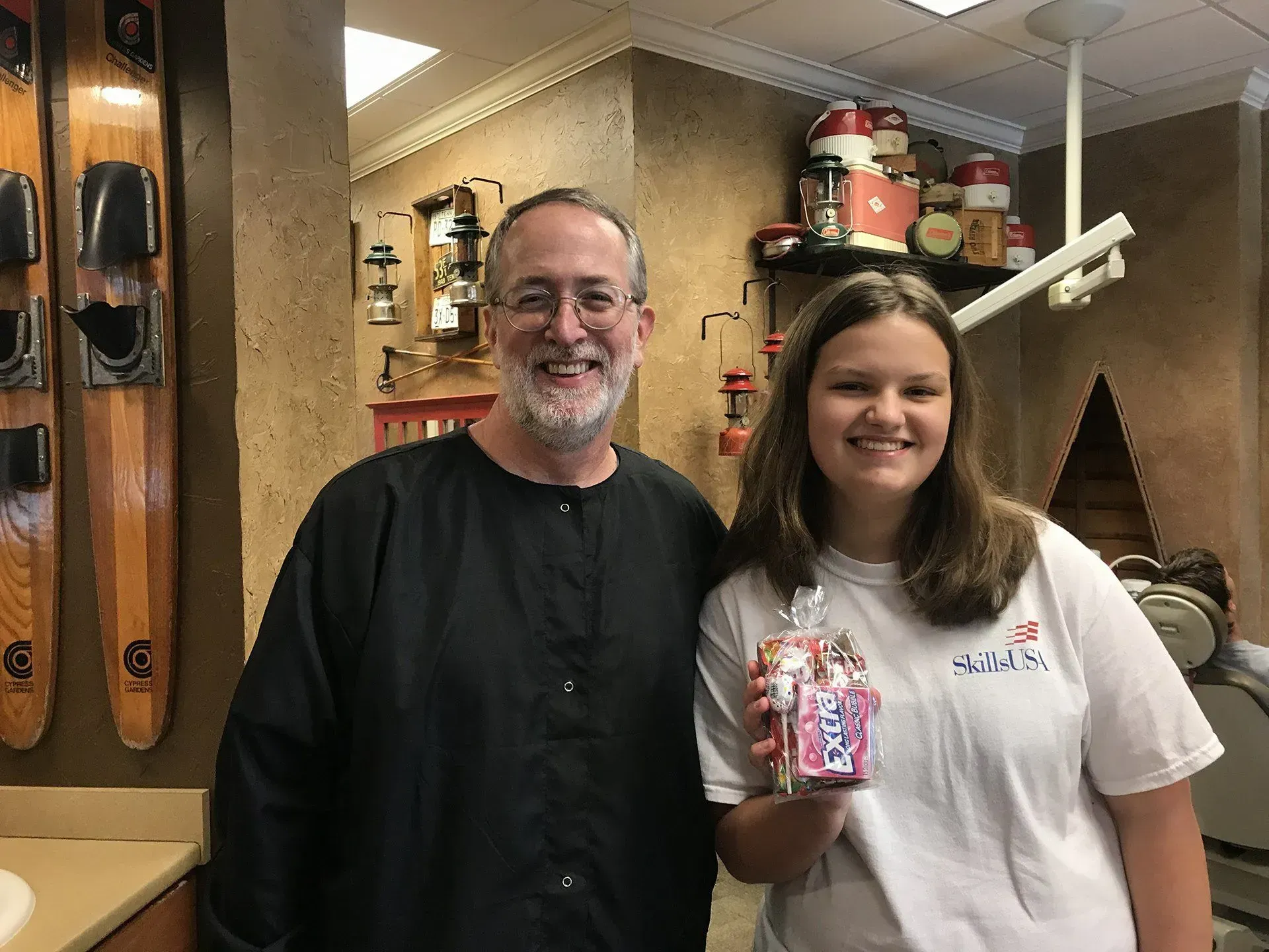 Man and woman smiling, holding gift in a dental office. Water skis and decorative items are visible.