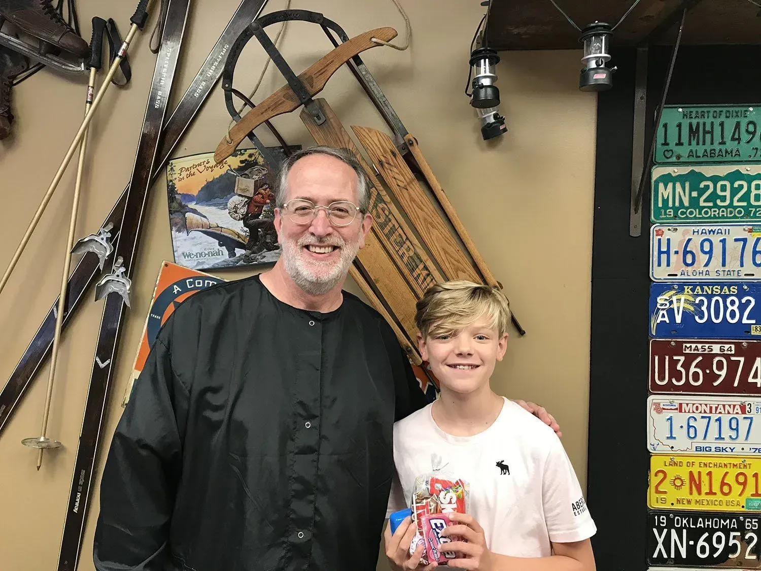 Man and boy stand together, smiling. The boy holds a box. Antique skis and license plates are in the background.