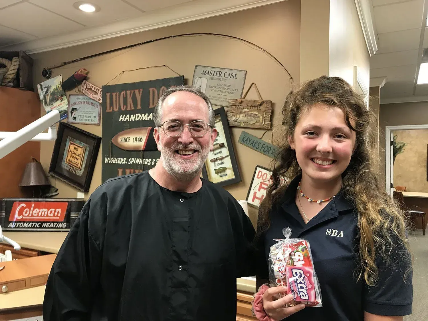 Dentist and patient smiling in office; patient holds candy; signage on wall.