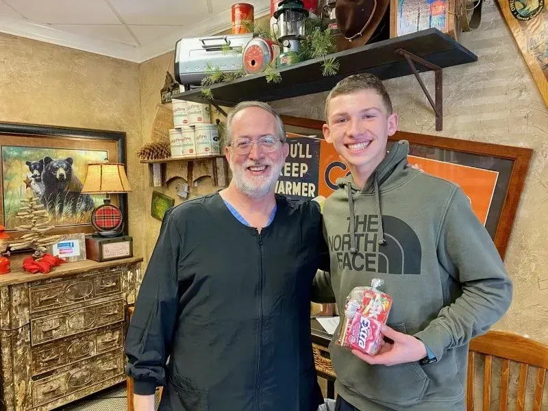 Man in scrubs and young man smiling, holding a gift, inside a rustic-themed office.