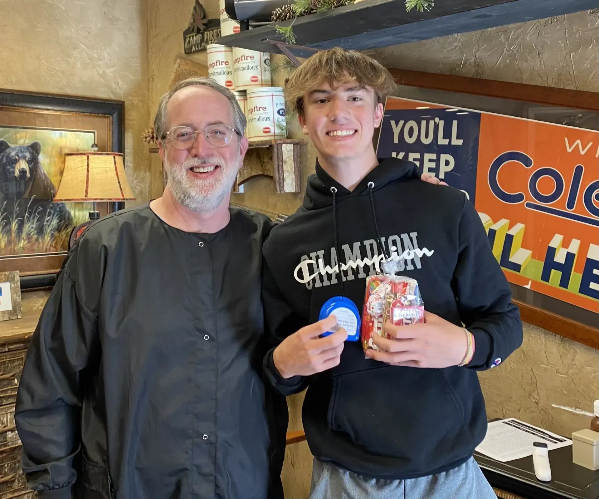 Two men stand together, smiling. One holds a can with a Christmas decoration. The background has shelves and signs.