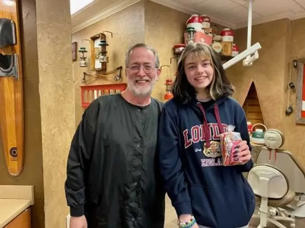 Man and girl smiling in a dental office. Girl holds a gift bag. Background has dental equipment and decor.