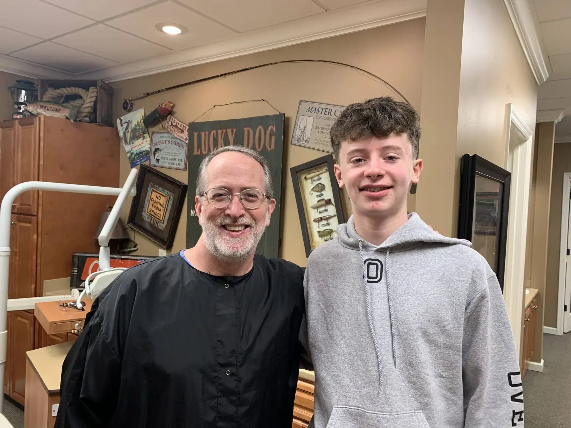 Man and teenager smiling in a dental office. The man wears a black cape. The teenager has braces.