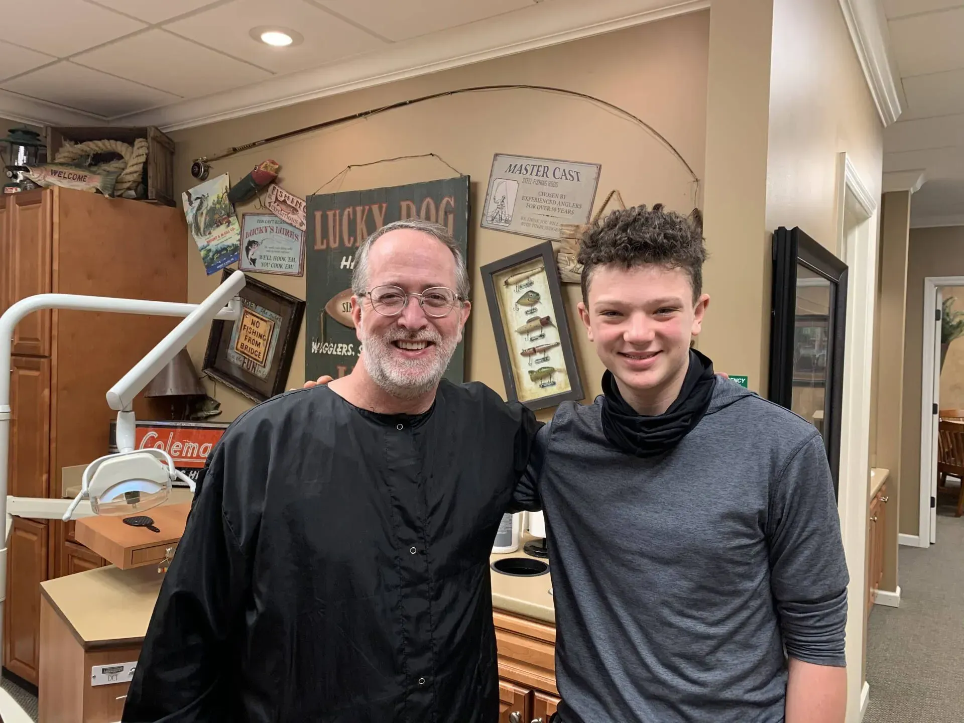 Man and teen pose together in dental office; smiling. Brown walls with decor visible.