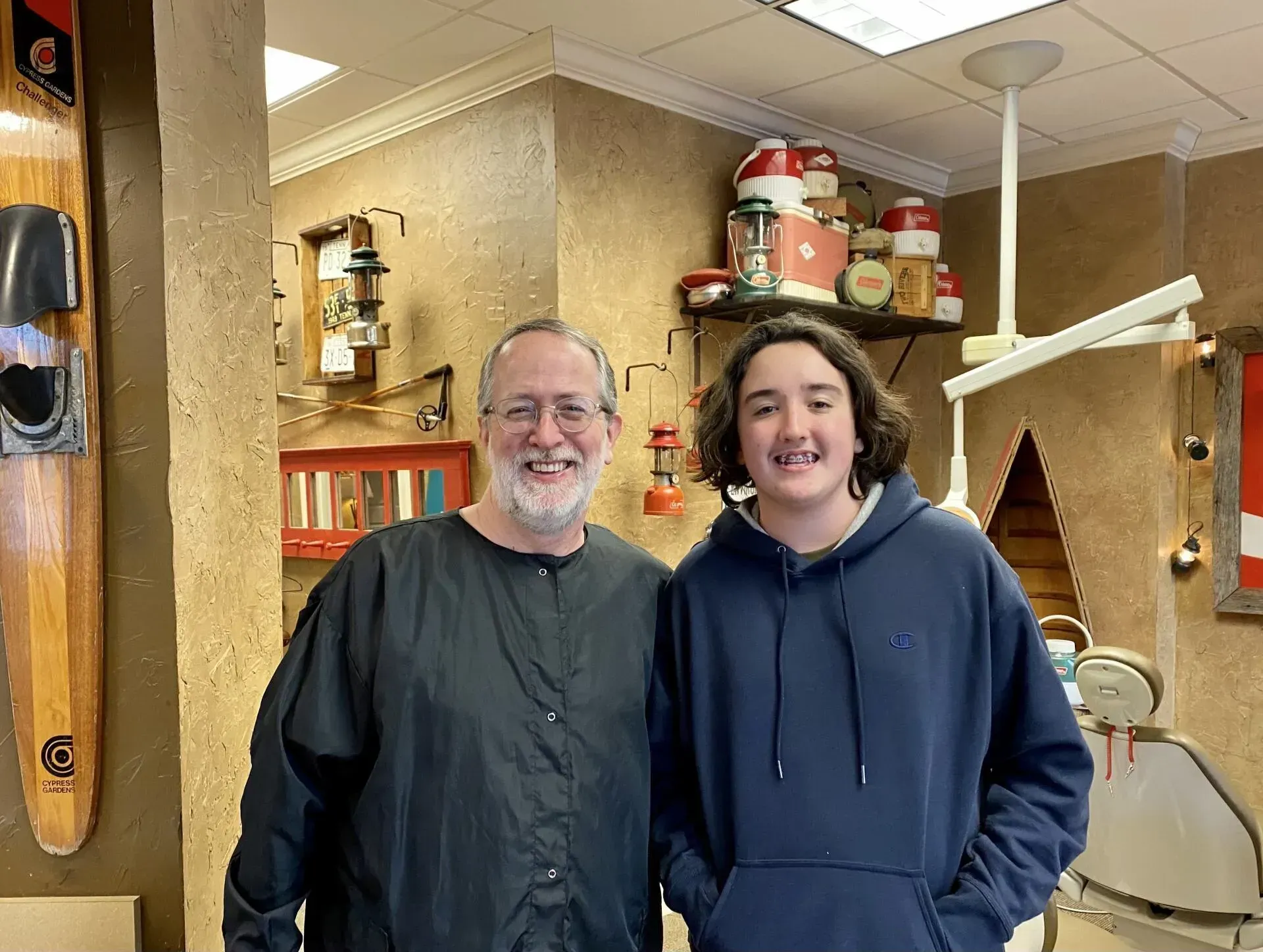Man and teen boy standing indoors, possibly a dentist's office, smiling.