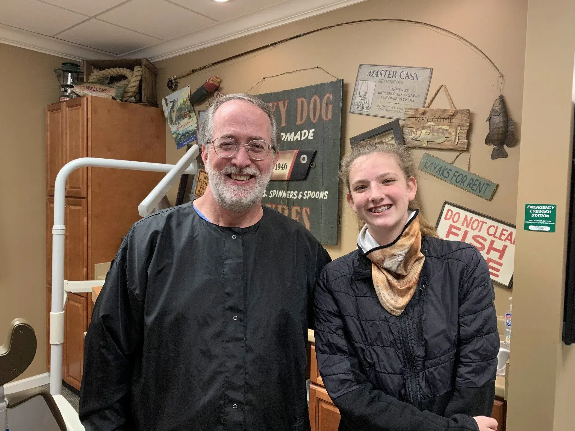 A smiling man and woman stand in a dental office. The man wears a black smock. The woman wears a black jacket.