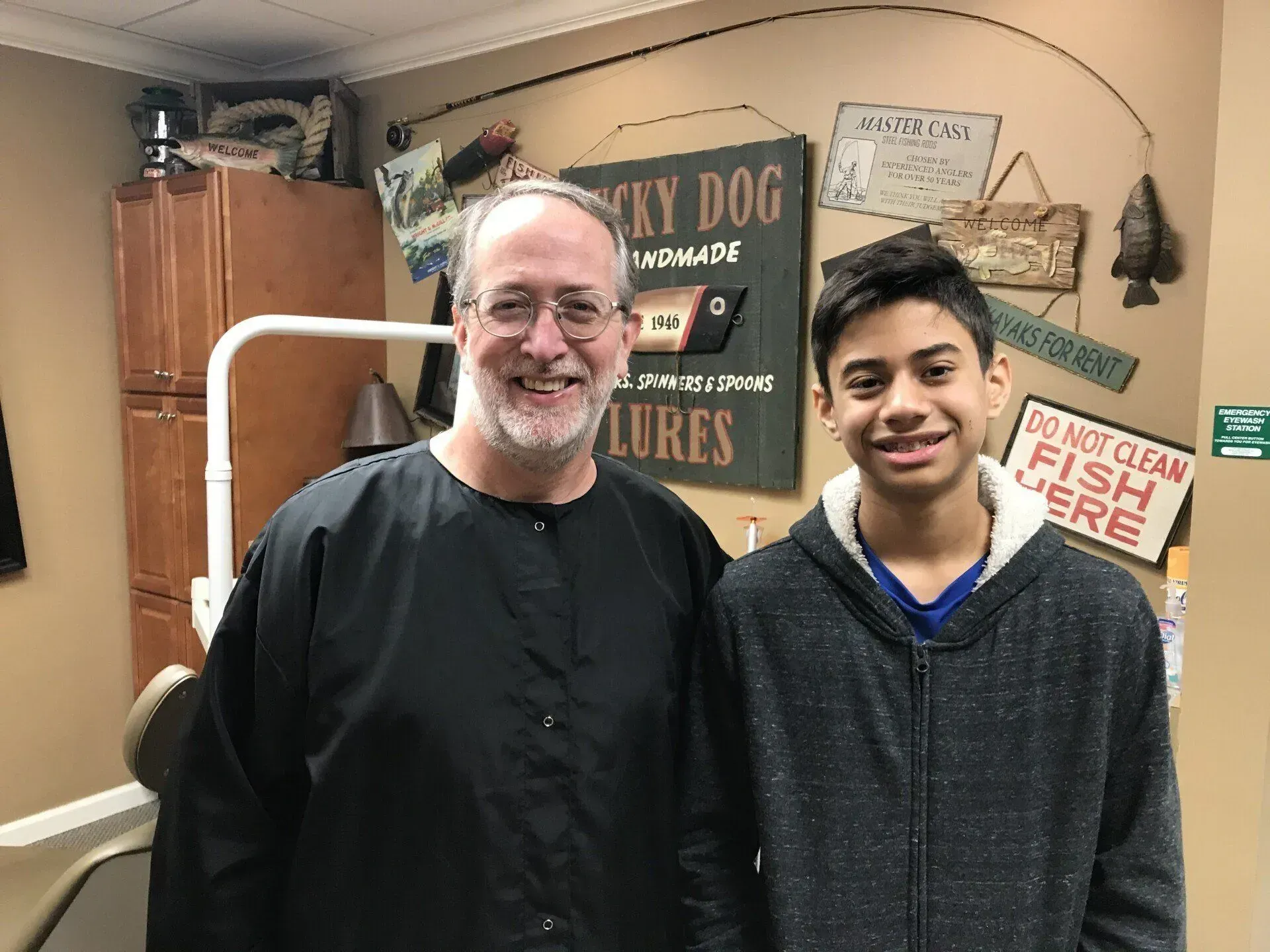 Man in dental garb and boy with braces smile together in dental office. Fishing-themed decor.