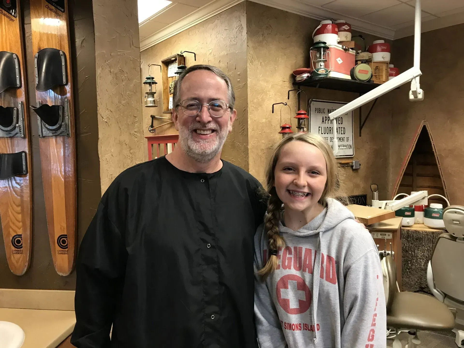 A dentist and a smiling girl posing together in a dental office. Water skis hang on the wall.